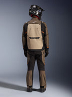 Motorista visto desde atrás con la chaqueta Alpinestars Cusco Drystar® en Walnut Aluminum Black, con un gran bolsillo trasero color aluminio y detalles en naranja, pantalones adventure a juego, casco off-road negro y guantes, equipamiento textil resistente para touring.