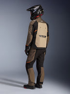 Chaqueta Alpinestars Cusco Drystar®, chaqueta de turismo de aventura, Walnut Aluminum Black, vista posterior que muestra el panel trasero en color canela y detalles en naranja, combinada con pantalones a juego y casco de fibra de carbono negro.