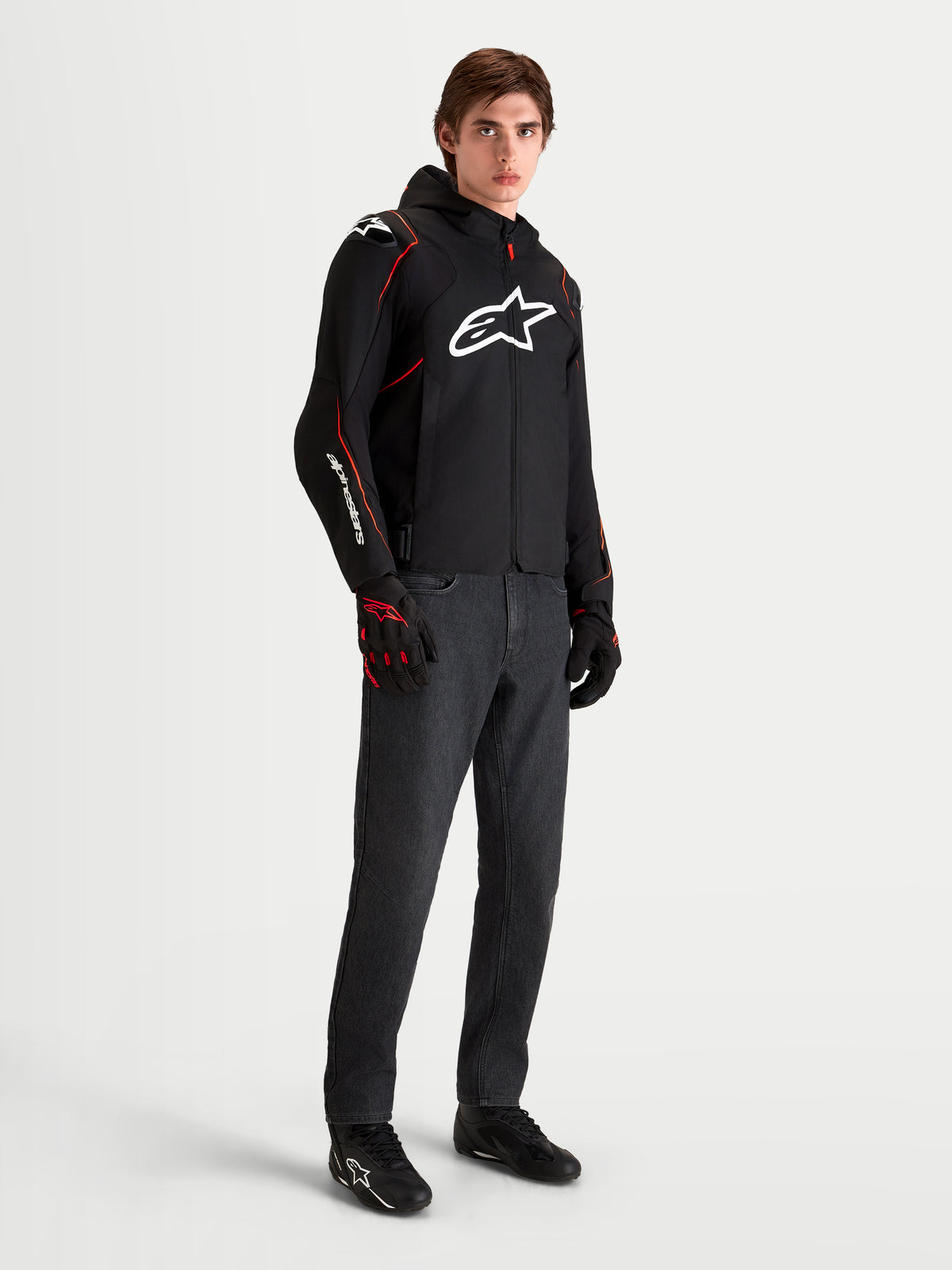 Alpinestars Zaca V2 Waterproof Jacket, chaqueta deportiva de moto para hombre negra y roja con capucha y logo blanco en el pecho, llevada por un modelo masculino con guantes de moto a juego y vaqueros oscuros
