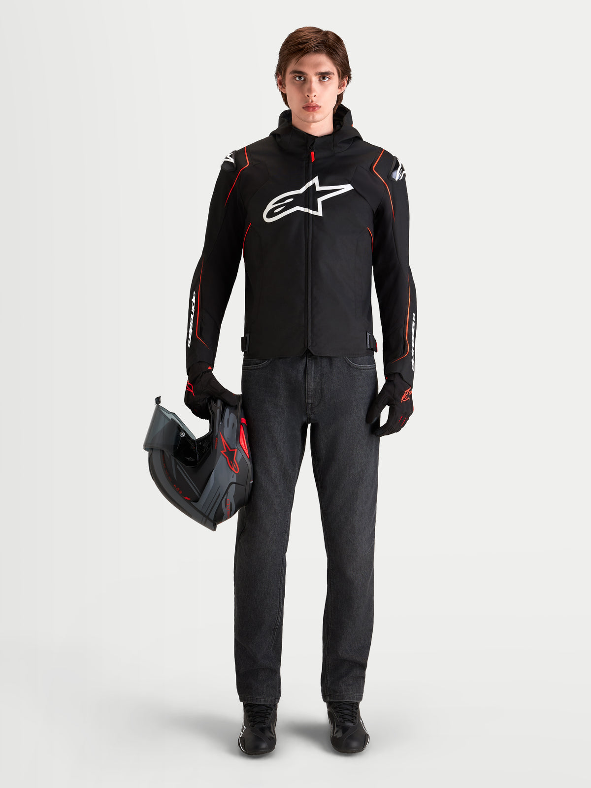 Chaqueta de moto Alpinestars Zaca V2 Waterproof, chaqueta deportiva de hombre negra y roja con protección en los hombros DFS y capucha integrada, vestida por un modelo que sostiene un casco