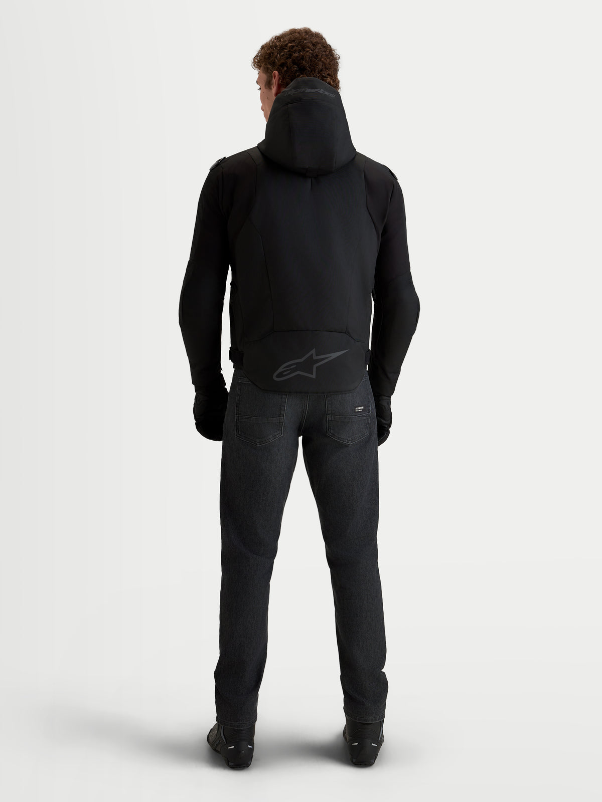 Alpinestars Zaca V2 Waterproof Jacket, chaqueta de moto negra para hombre vista desde la espalda con capucha integrada, dobladillo trasero extendido con logo tonal y equipo de protección de conducción