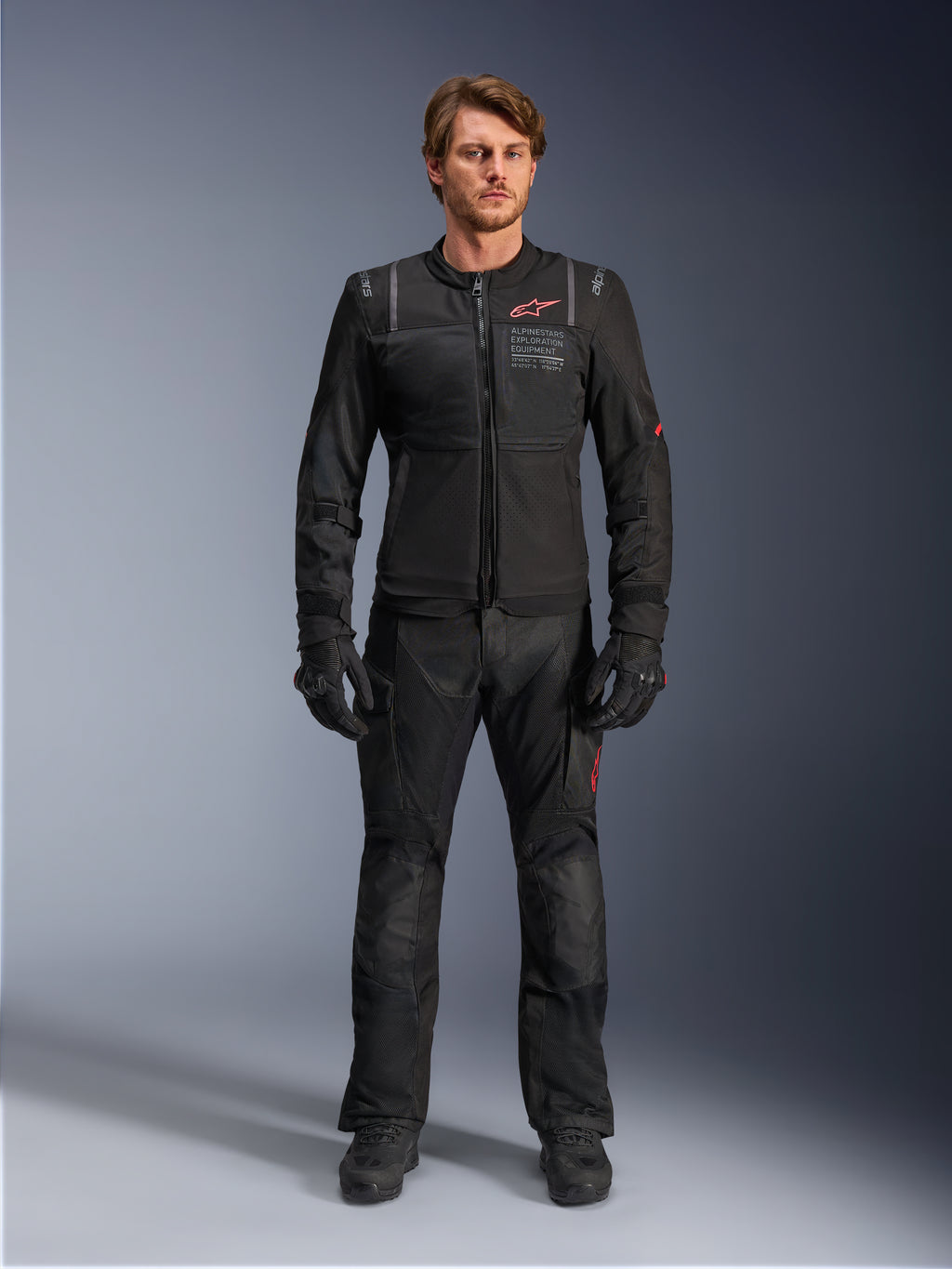 Male model wearing Alpinestars ST-2 Air Jacket, Black motorcycle jacket with mesh panels and red logo, 'Alpinestars Exploration Equipment' text on chest, paired with black technical riding pants and gloves, modern adventure touring gear