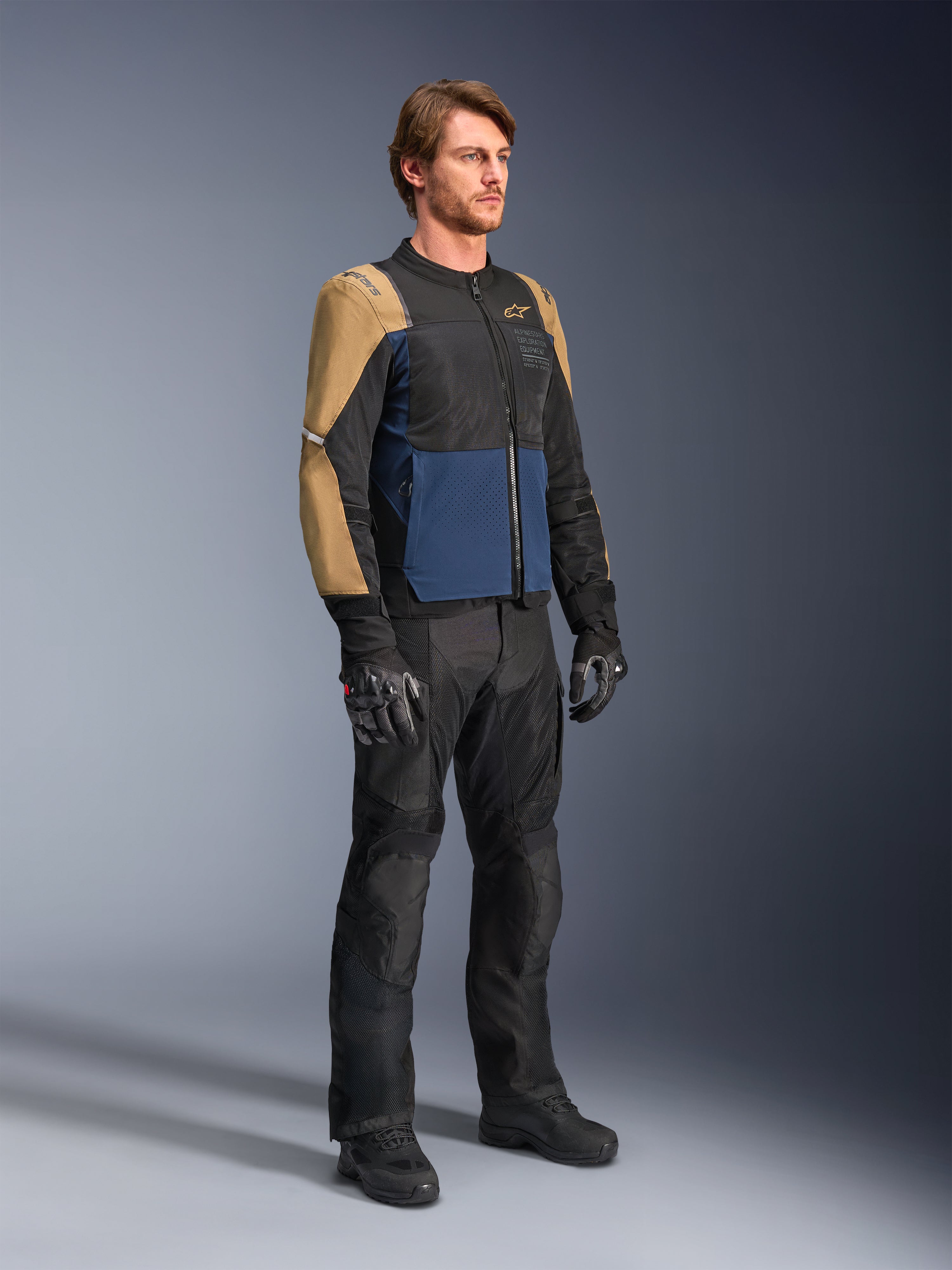 Piloto con la chaqueta Alpinestars ST-2 Air en azul profundo, caqui oscuro y negro, con grandes paneles de malla y perforaciones láser, combinada con pantalones de motociclista, guantes y botas negras para un look completo de turismo de verano.