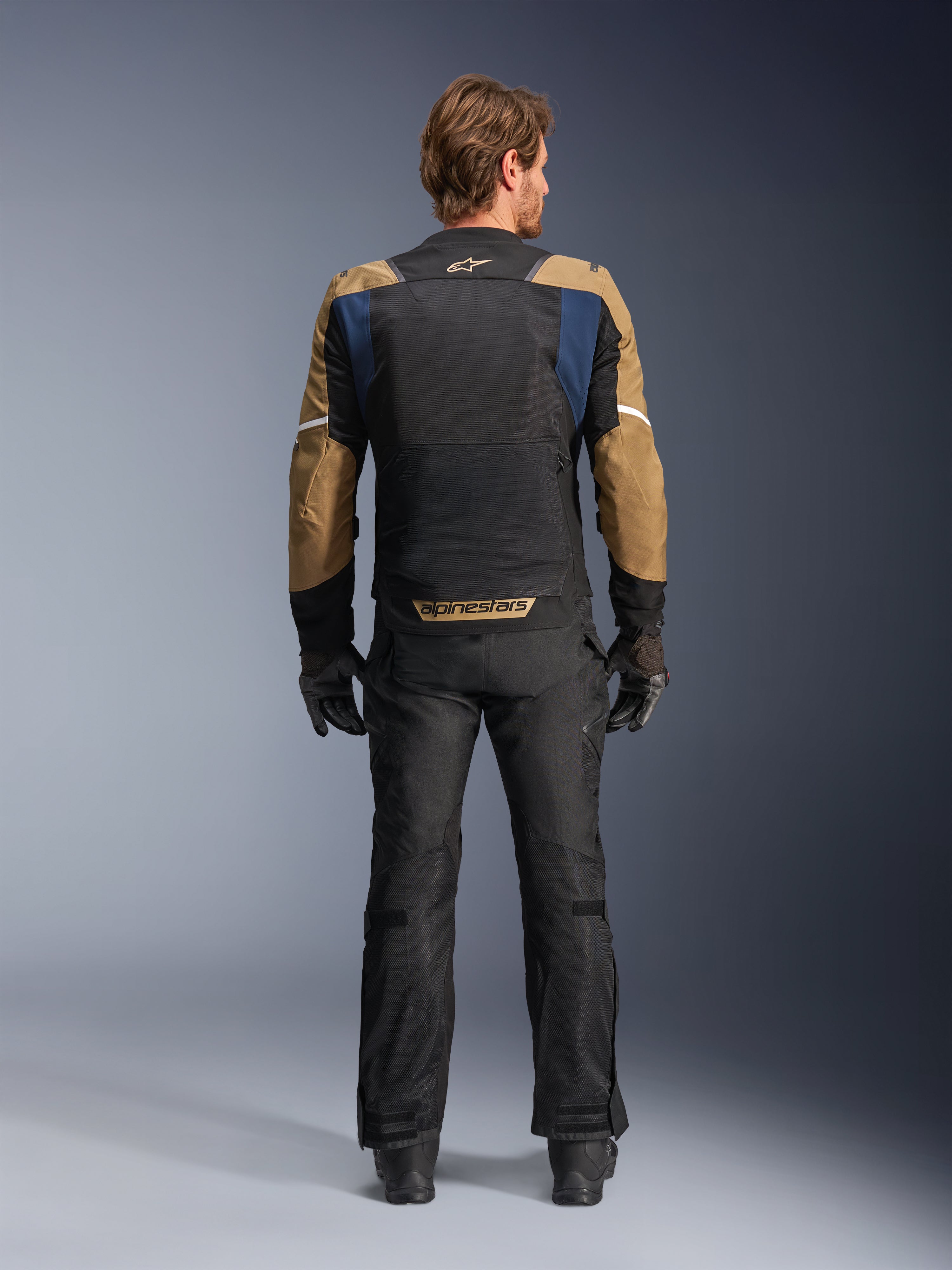 Chaqueta Alpinestars ST-2 Air en azul profundo, caqui oscuro y negro, vista trasera en modelo que muestra el gran panel de malla en la espalda y mangas caqui con rayas blancas, combinada con pantalones técnicos de moto y guantes negros para turismo de aventura.