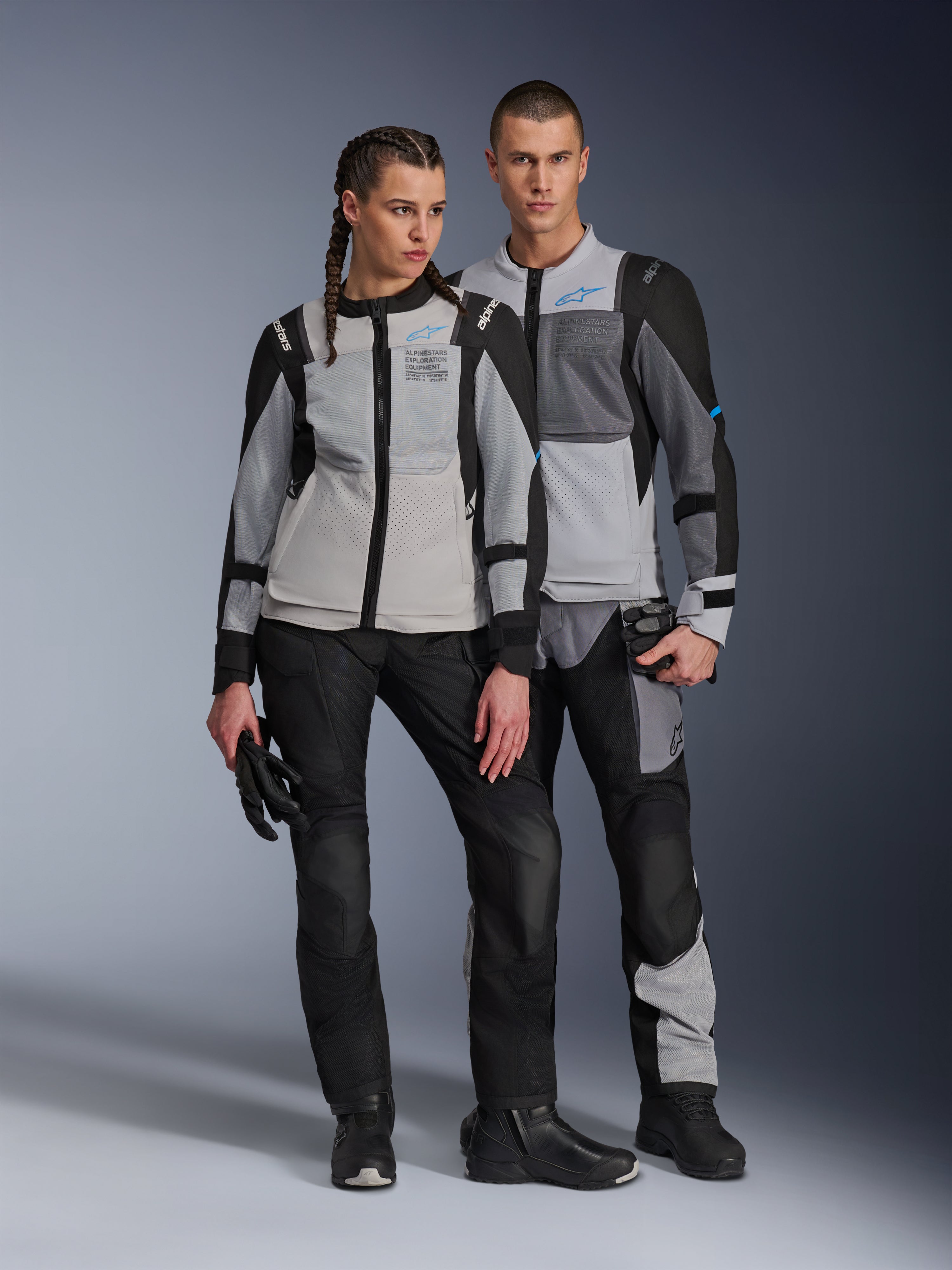 Modelos masculino y femenino luciendo chaquetas Alpinestars ST-2 Air en Ice Gray, Dark Gray y Black, equipamiento técnico de motociclismo para touring de verano con amplios paneles de malla y marca Exploration Equipment, combinados con pantalones de pilotaje, guantes y botas negras.