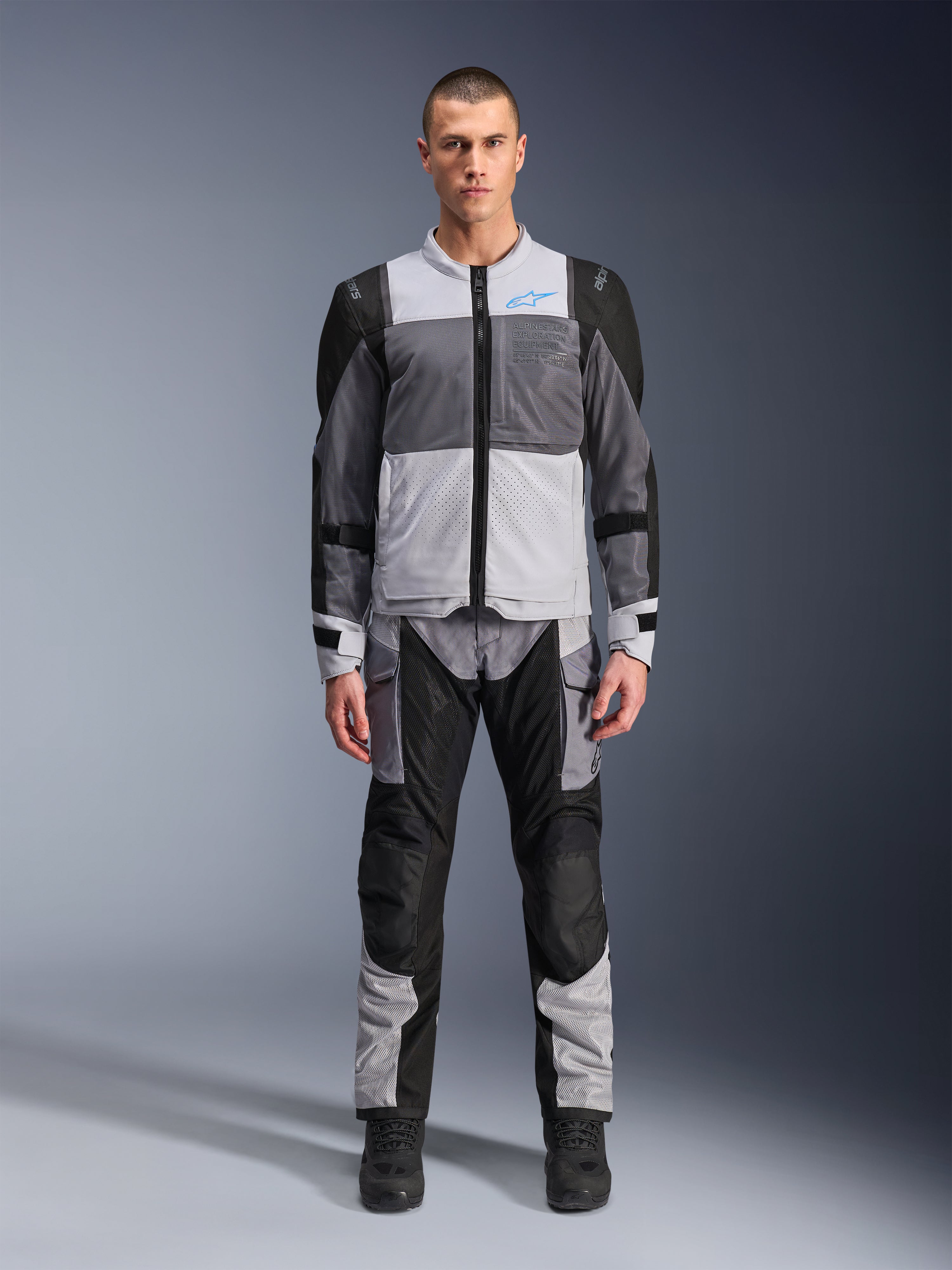 Chaqueta Alpinestars ST-2 Air, chaqueta de touring para moto, en gris hielo, gris oscuro y negro, vestida por un modelo con pantalones de aventura a juego, con grandes paneles de malla, logotipo de Alpinestars azul en el pecho y marca Alpinestars Exploration Equipment.