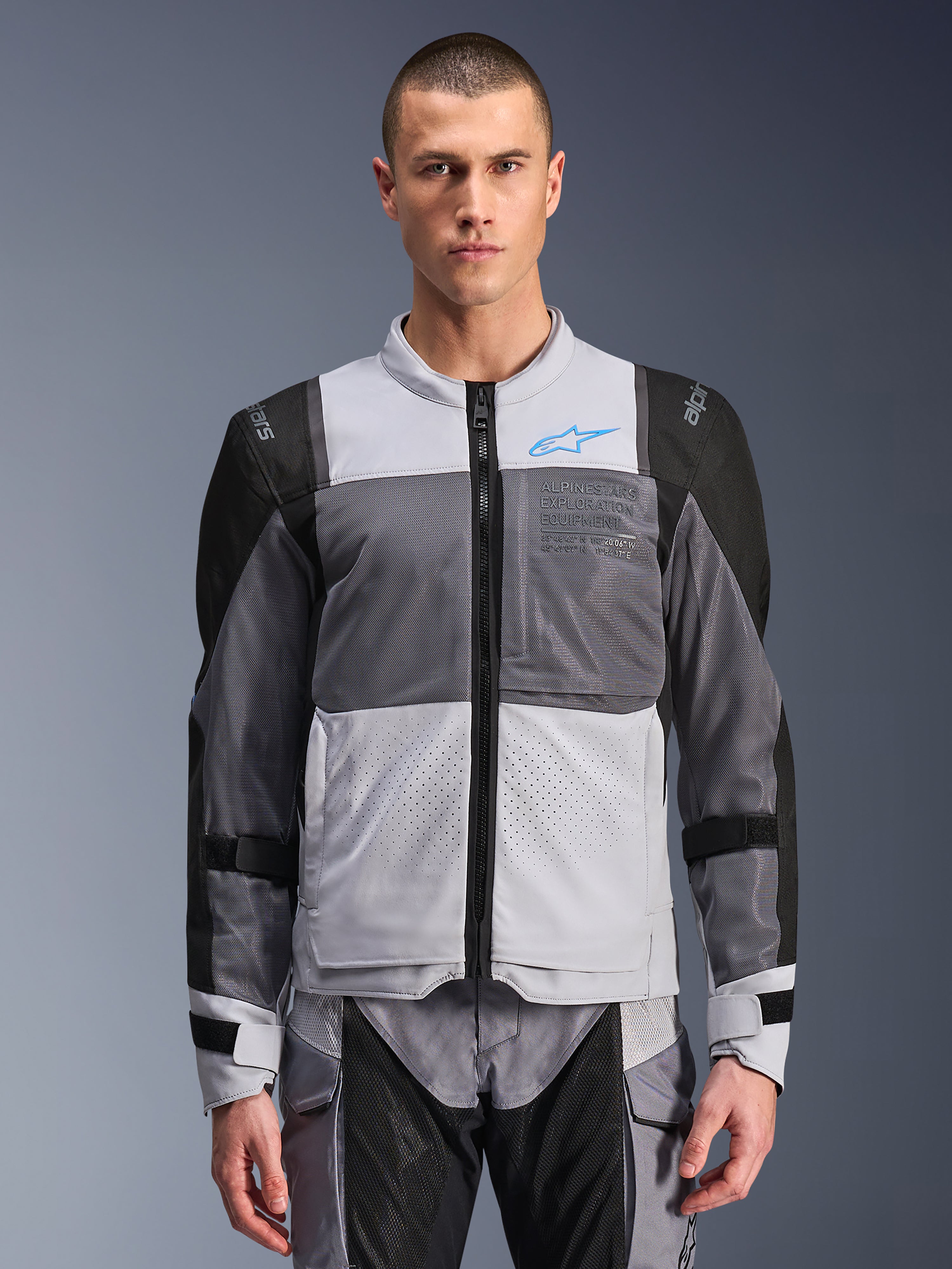 Chaqueta Alpinestars ST-2 Air, chaqueta de moto para hombre, gris hielo y gris oscuro con detalles en negro, amplios paneles de ventilación de malla, frontal perforado con láser, logotipo azul de Alpinestars, cremallera frontal, usada por un modelo con pantalones de moto a juego
