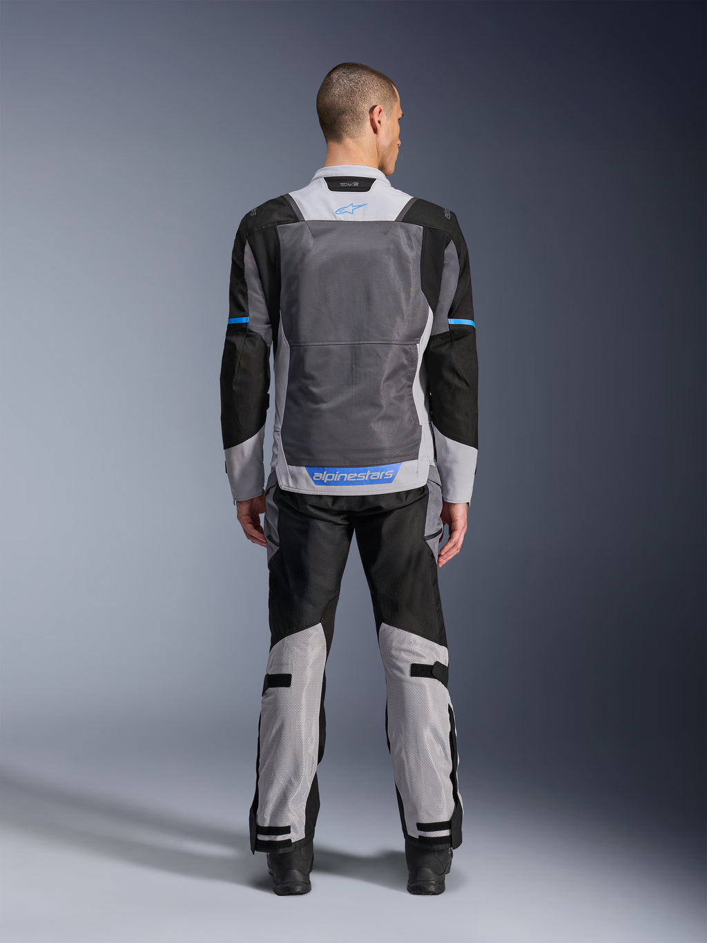 Chaqueta Alpinestars ST-2 Air, Ice Gray Dark Gray Black, vista trasera en modelo, con amplios paneles de ventilación de malla, detalles azules y logotipo de Alpinestars, combinada con pantalones de moto a juego para turismo de aventura.