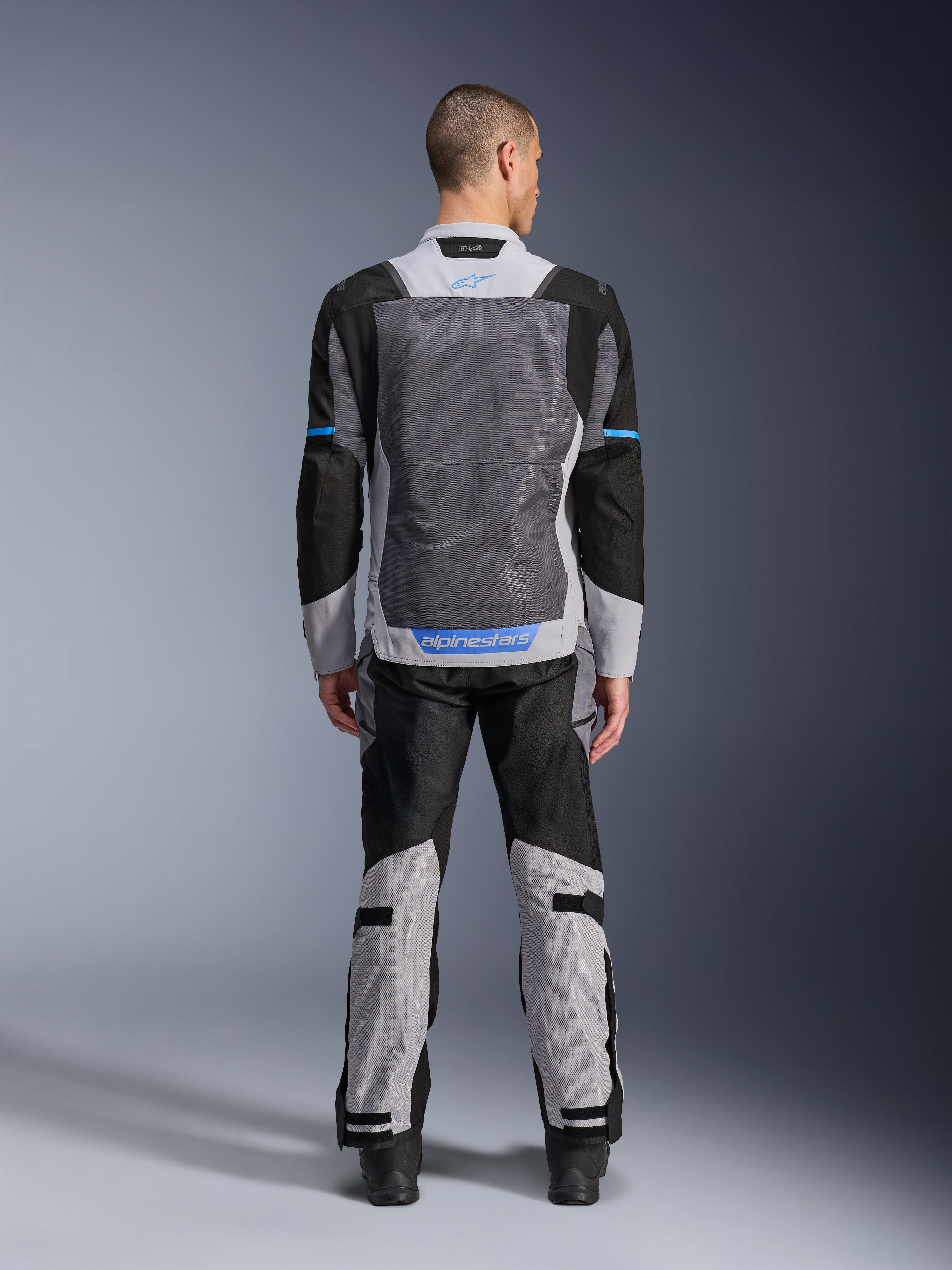 Chaqueta Alpinestars ST-2 Air, Ice Gray Dark Gray Black, vista trasera en modelo, con amplios paneles de ventilación de malla, detalles azules y logotipo de Alpinestars, combinada con pantalones de moto a juego para turismo de aventura.