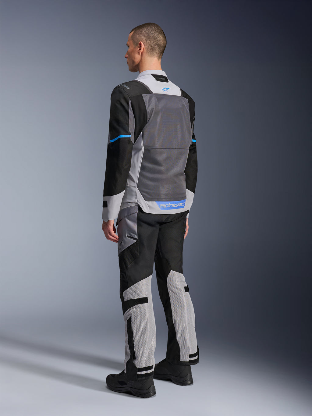 Modelo masculino de espaldas luciendo la chaqueta Alpinestars ST-2 Air en gris hielo, gris oscuro y negro, con un gran panel de malla gris en la espalda, detalles del logo en azul y pantalones de moto a juego para turismo de verano.