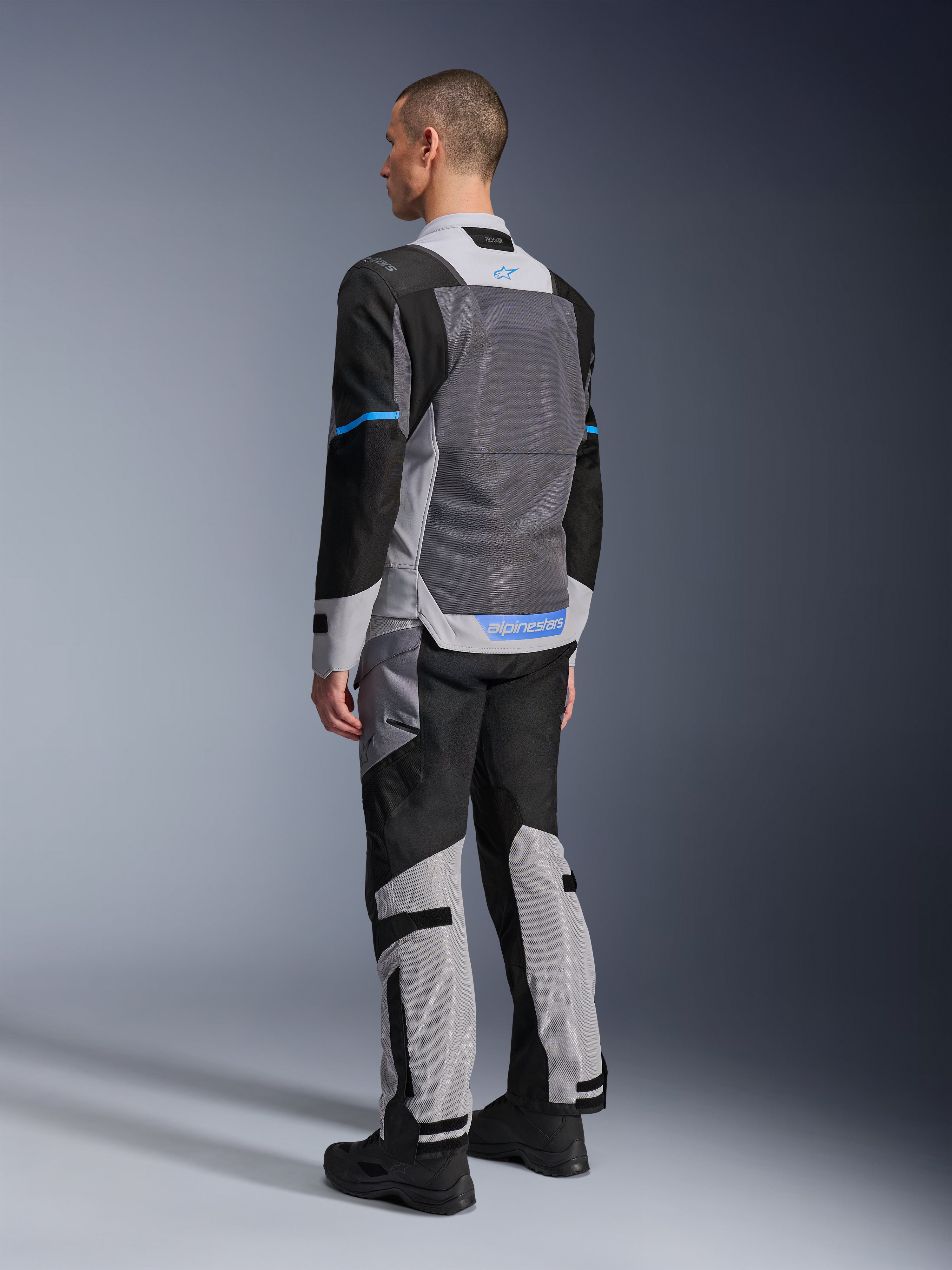 Modelo masculino de espaldas luciendo la chaqueta Alpinestars ST-2 Air en gris hielo, gris oscuro y negro, con un gran panel de malla gris en la espalda, detalles del logo en azul y pantalones de moto a juego para turismo de verano.