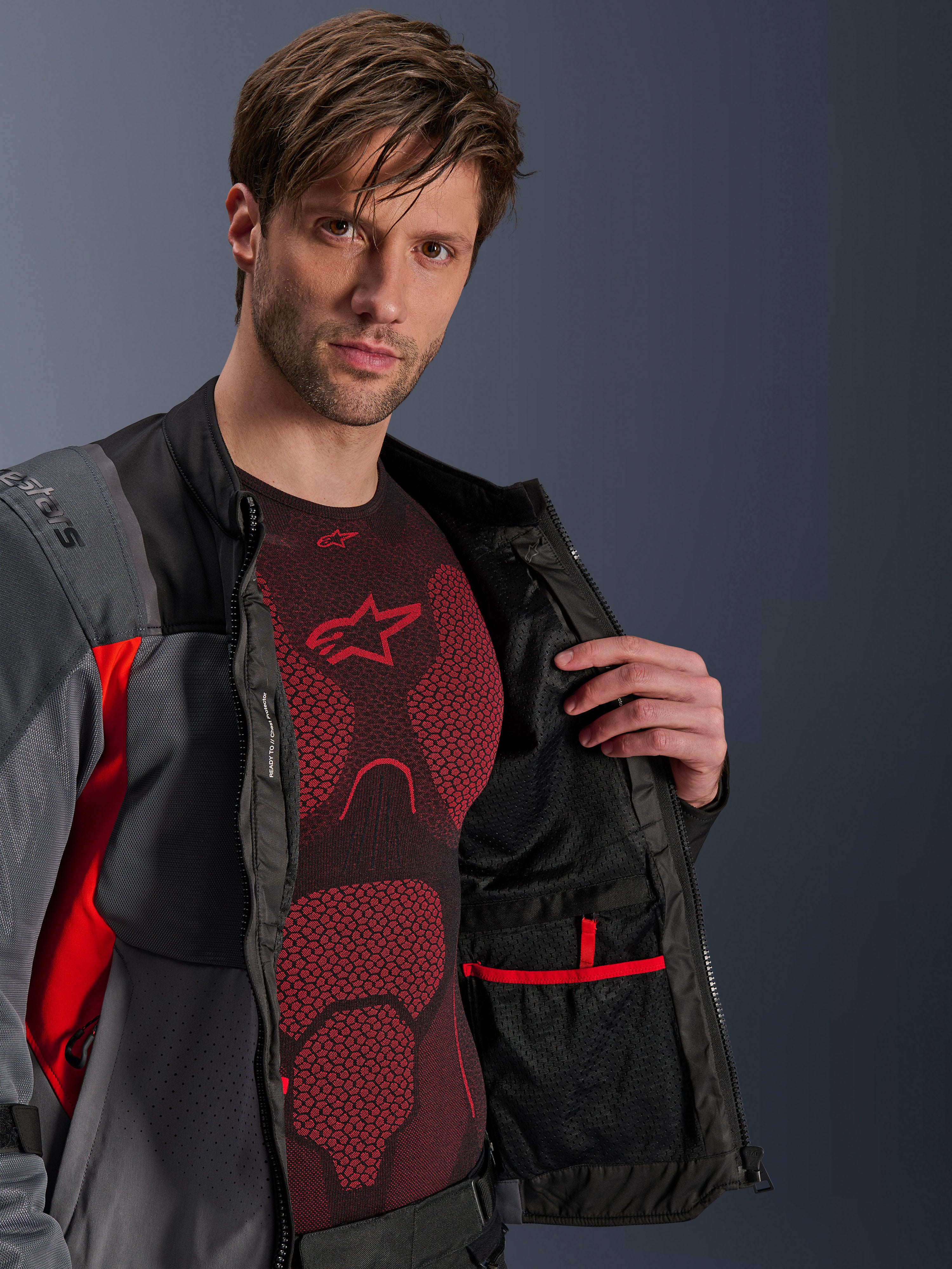 Alpinestars ST-2 Air Jacket, Deep Gray Dark Gray and Bright Red, unzipped to show internal mesh lining and red-trimmed pocket, worn over a red technical base layer, modern adventure motorcycle touring gear