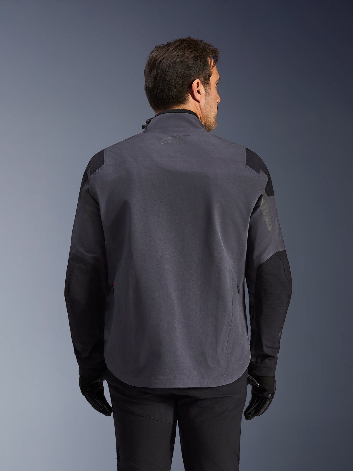 Alpinestars Maxdura Drystar®XF Laminated Armored Shell, chaqueta técnica de aventura, gris oscuro y negro, vista trasera con paneles reforzados en hombros y codos, sutil logo en la parte superior de la espalda, usada con guantes y pantalones negros, diseñada para protección en todo tipo de clima