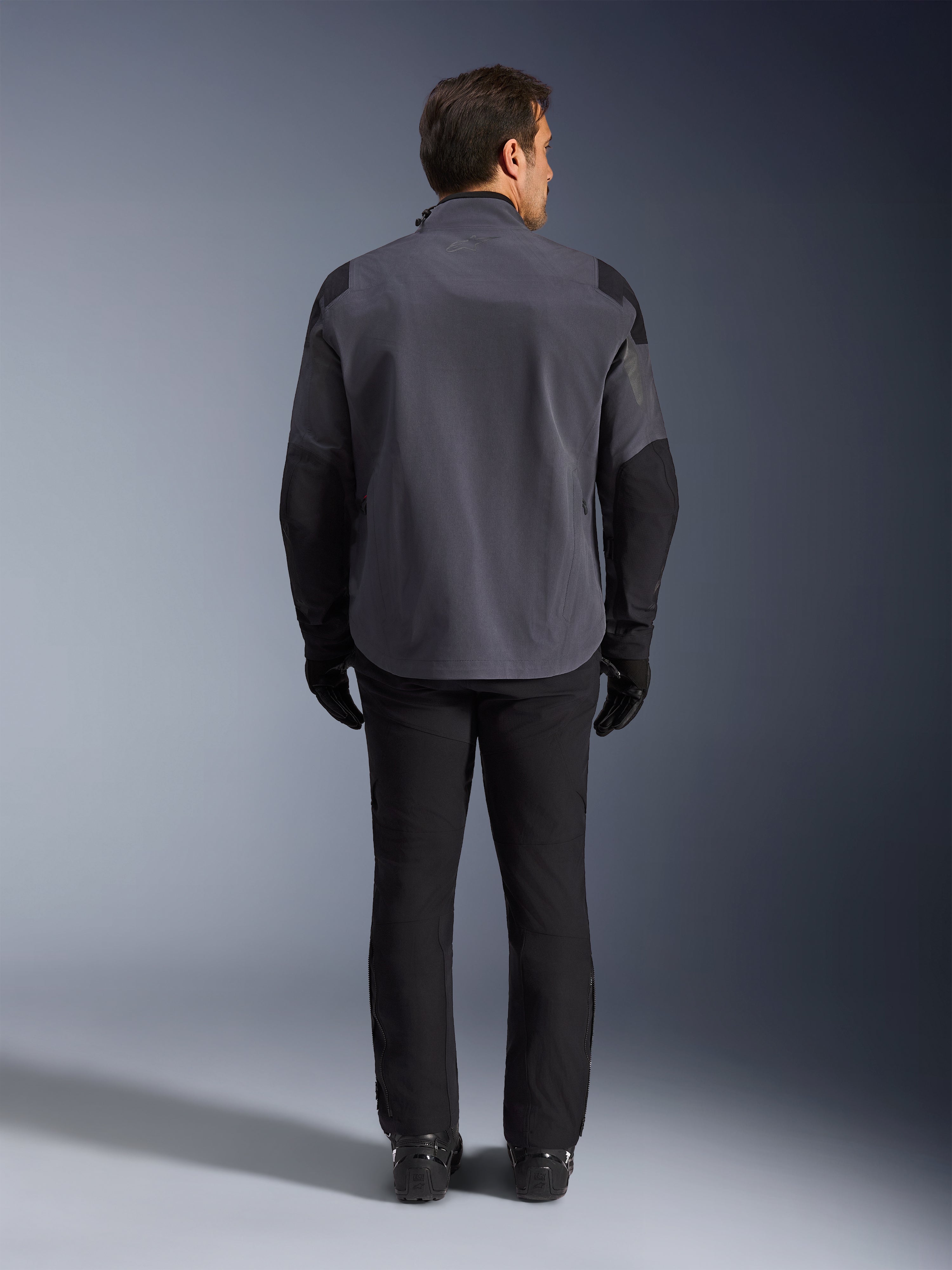 Alpinestars Maxdura Drystar®XF Laminated Armored Shell, chaqueta de motocicleta, gris oscuro y negro, vista trasera de un piloto con equipamiento completo de aventura, capa técnica laminada con paneles reforzados negros en hombros y codos, combinada con pantalones y guantes negros.