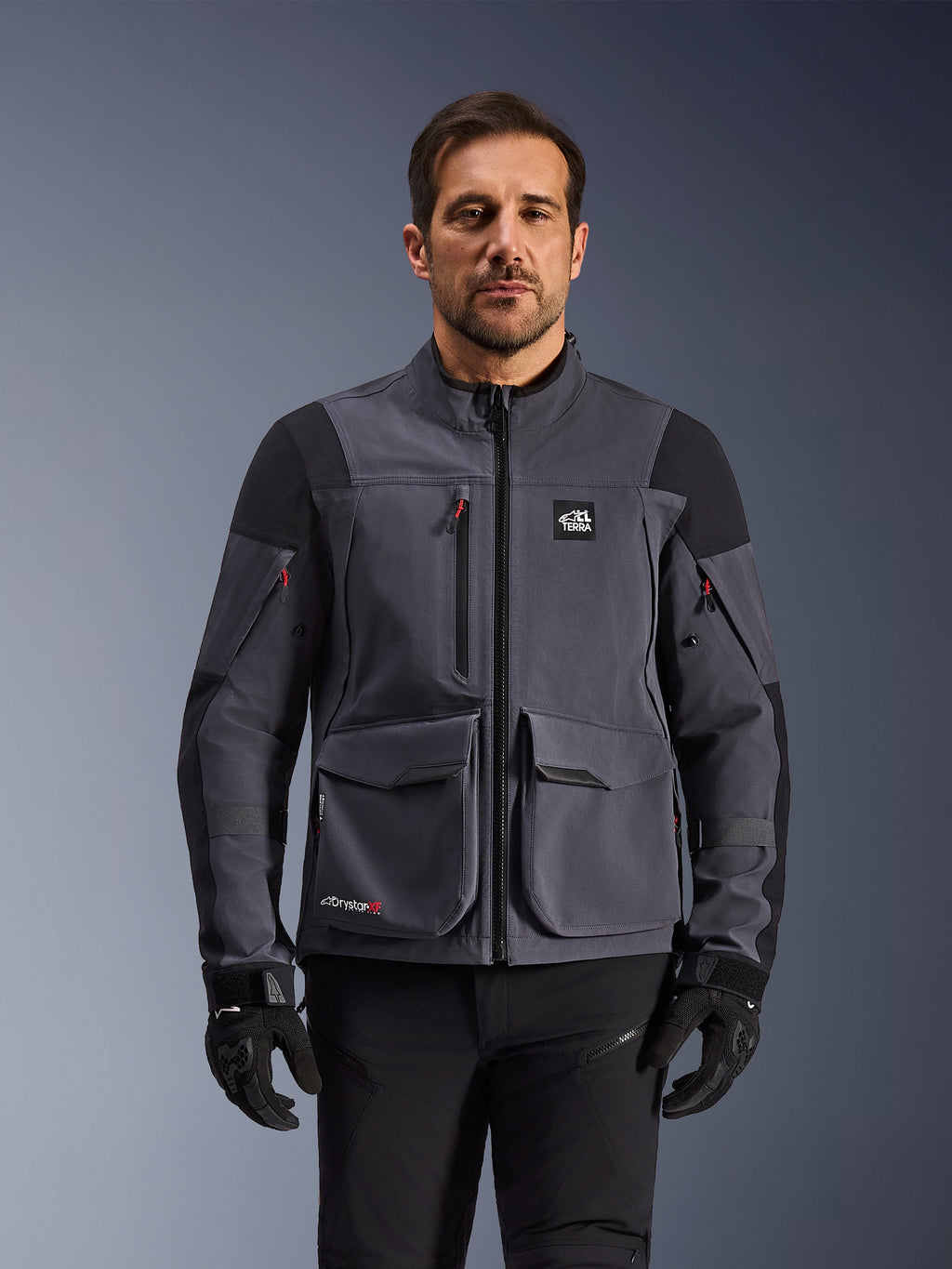 Alpinestars Maxdura Drystar®XF Laminated Armored Shell, chaqueta de moto de aventura en gris oscuro y negro, vista frontal en modelo, con grandes bolsillos de carga, logotipo 'All Terra' en el pecho y cremalleras de ventilación, combinada con guantes y pantalones de montar negros.