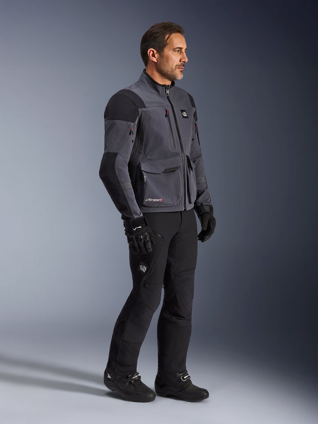 Alpinestars Maxdura Drystar®XF Laminated Armored Shell, chaqueta de motociclista en gris oscuro y negro, capa técnica dual-sport usada con pantalones y botas negras a juego, con bolsillos de carga y paneles reforzados en los hombros, foto de estudio de equipamiento completo para conducción de aventura