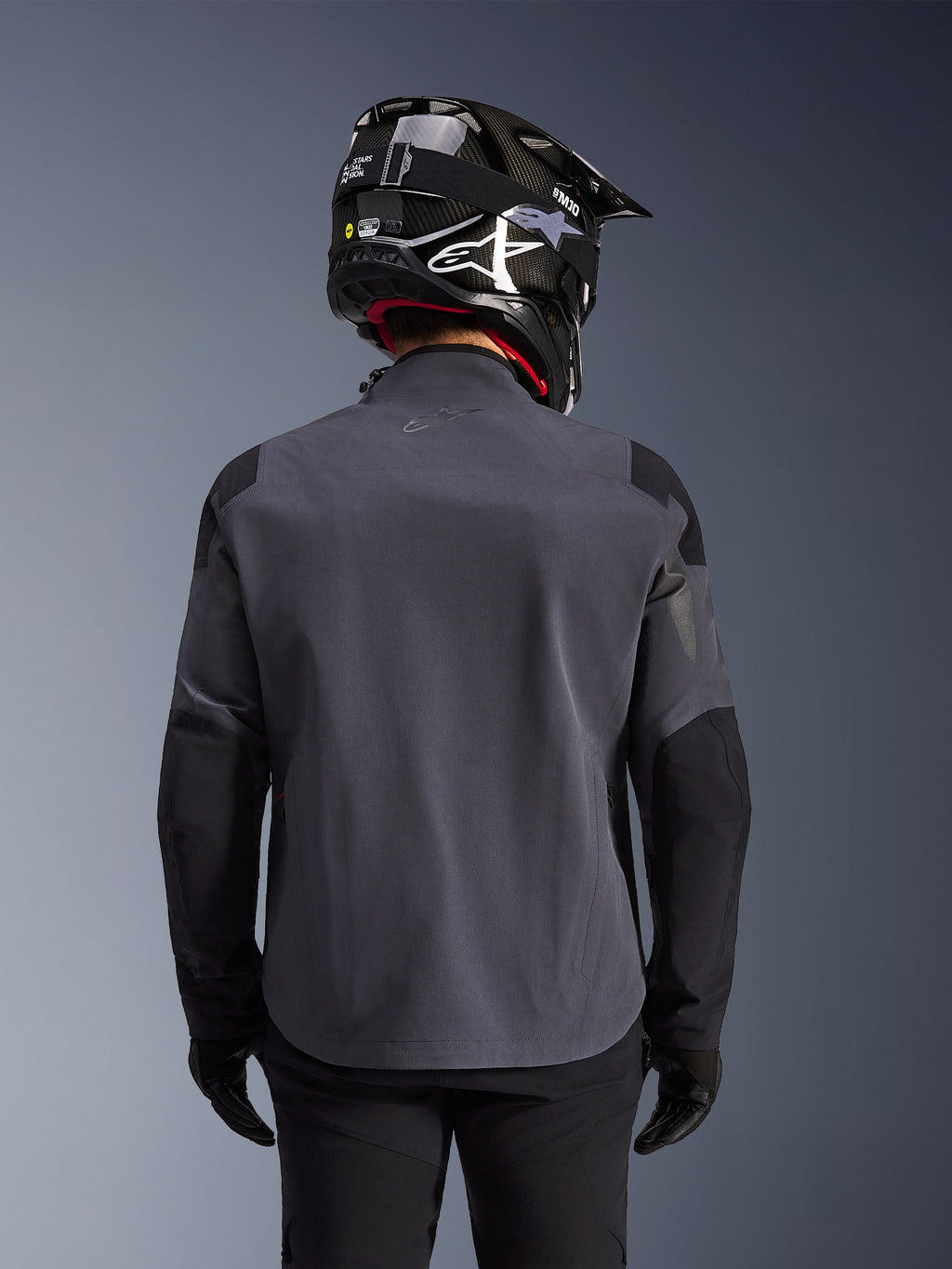 Maxdura Drystar®XF Laminated Armored Shell, chaqueta de moto de aventura, Deep Gray Black, vista posterior que muestra la construcción textil técnica y los paneles reforzados, usada con un casco de fibra de carbono negro, pantalones de montar negros y guantes