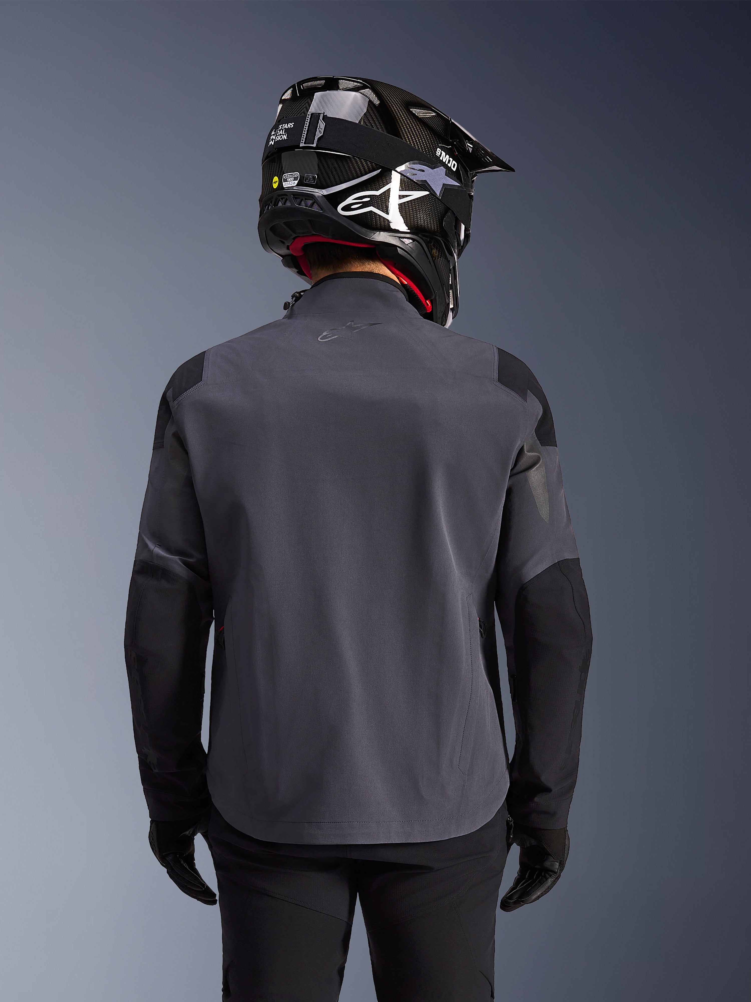 Maxdura Drystar®XF Laminated Armored Shell, chaqueta de moto de aventura, Deep Gray Black, vista posterior que muestra la construcción textil técnica y los paneles reforzados, usada con un casco de fibra de carbono negro, pantalones de montar negros y guantes