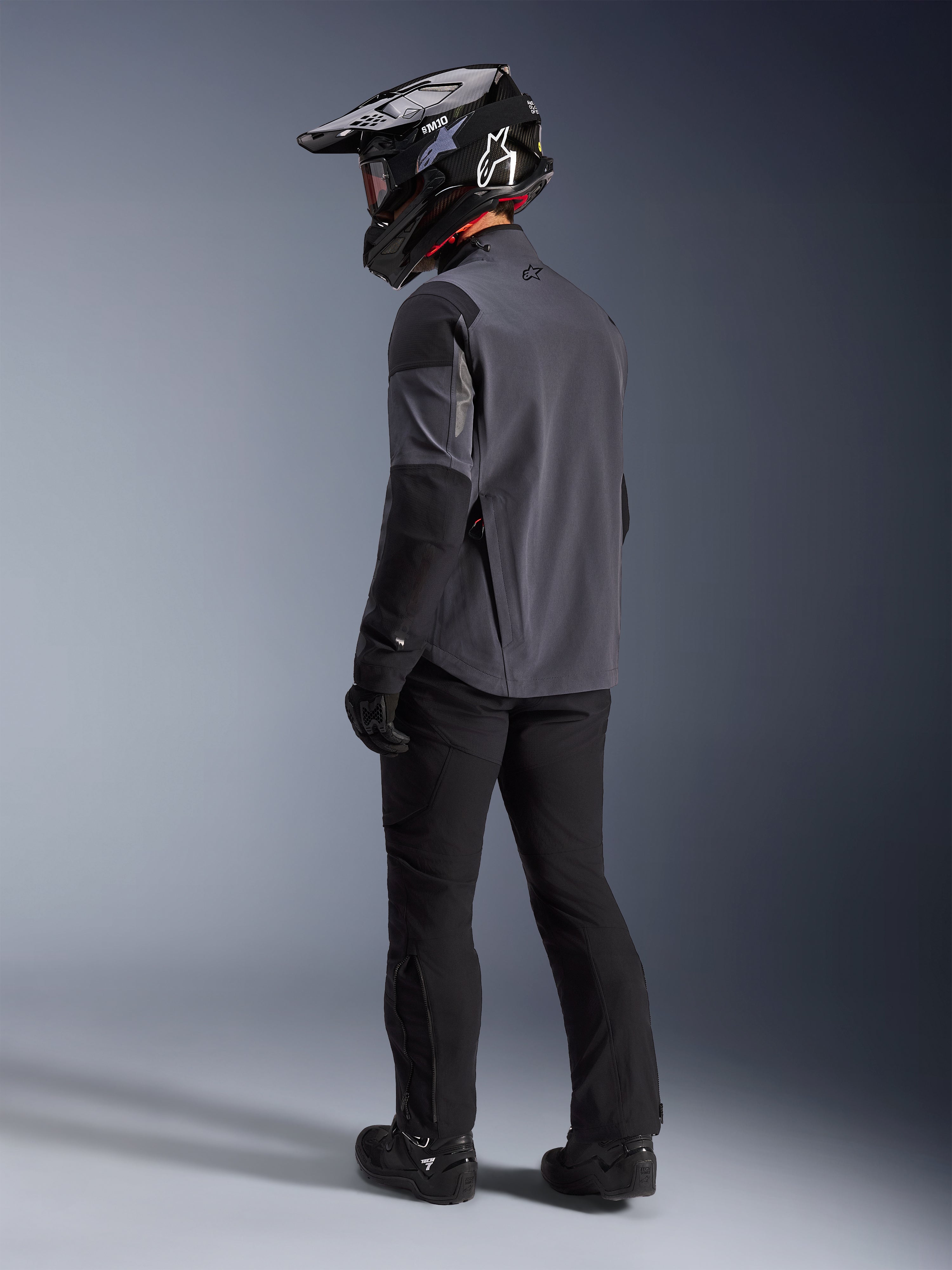 Motociclista de aventura con la chaqueta Maxdura Drystar®XF Laminada con Armadura en Deep Gray Black, vista posterior. Equipamiento completo con pantalones de moto negros, casco de motocross negro con marca Alpinestars y botas off-road negras. Conjunto técnico para todo clima para rendimiento dual-sport.