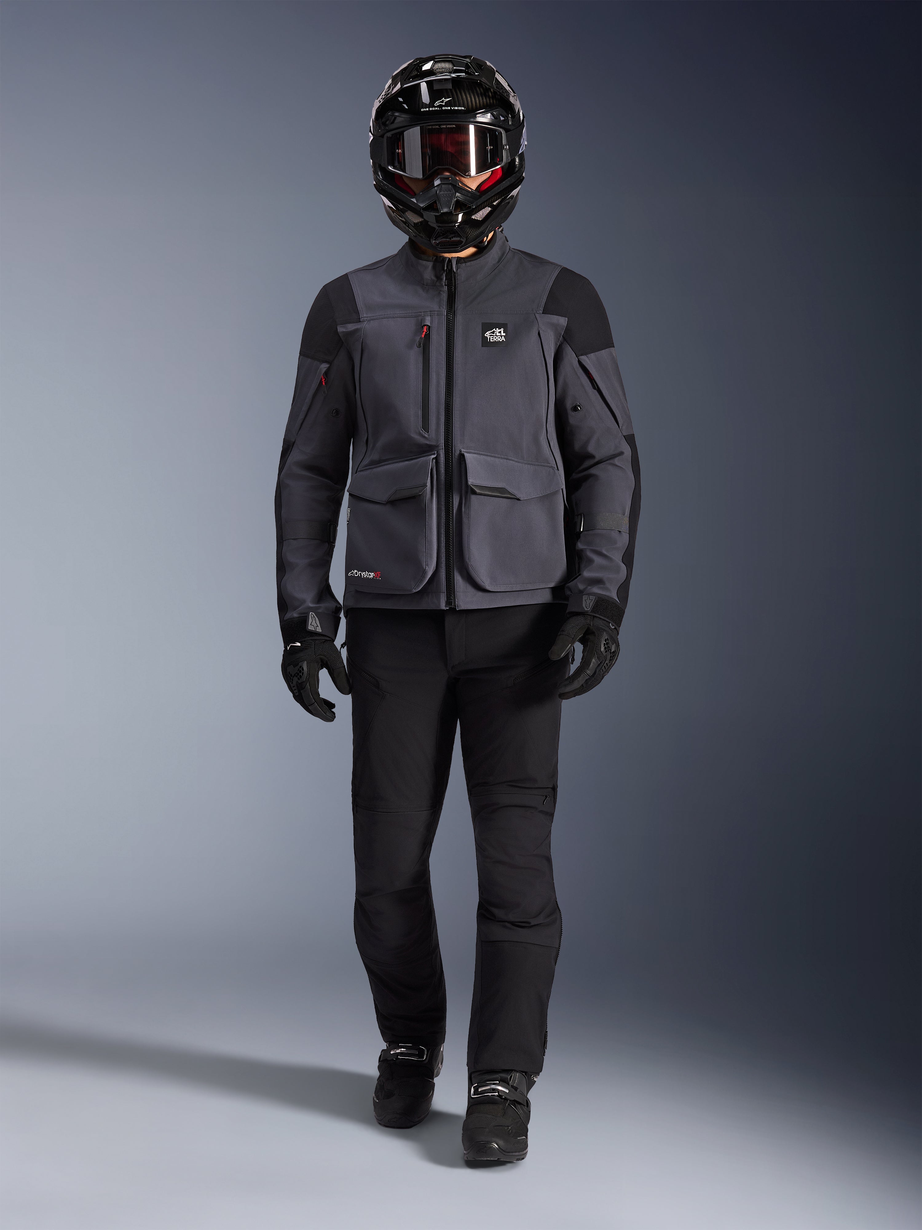 Alpinestars Maxdura Drystar®XF Laminated Armored Shell, chaqueta de motociclismo de aventura en gris oscuro y negro, usada con pantalones de montar negros a juego, un casco integral negro, guantes y botas, con bolsillos de carga prominentes y marca ALL TERRA para un estilo técnico de touring todoterreno
