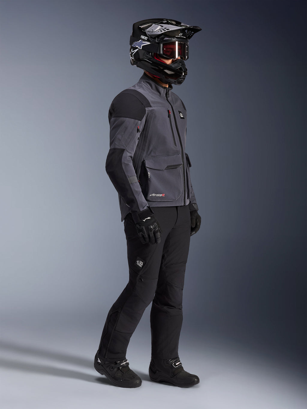 Motociclista con chaqueta Alpinestars Maxdura Drystar®XF Laminated Armored Shell en gris oscuro y negro, combinada con pantalones de aventura negros, casco negro con gafas, guantes y botas negros, toma de estudio de cuerpo completo que destaca el equipamiento técnico dual-sport.