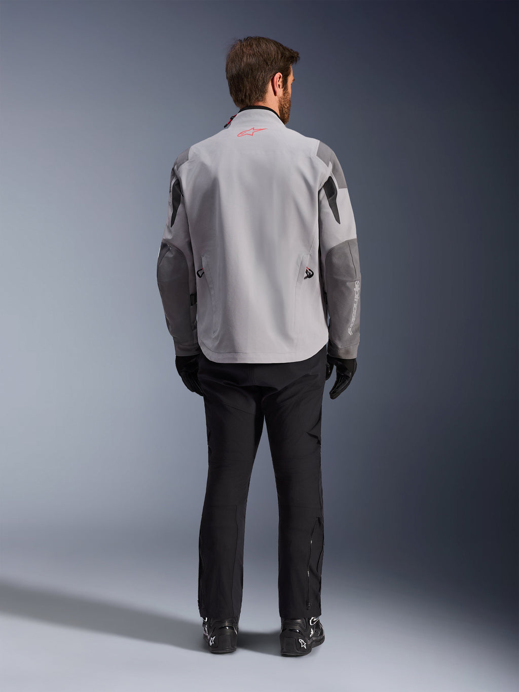 Alpinestars Maxdura Drystar®XF Laminated Armored Shell, chaqueta de motociclismo en gris medio y gris oscuro, vista trasera, con un cuerpo gris claro, paneles gris oscuro en los hombros y las mangas, un pequeño logotipo de la estrella Alpinestars en rojo en la parte posterior del cuello y marca blanca en la manga izquierda, combinada con pantalones y botas de montar negros.