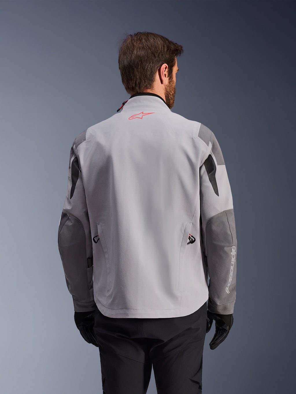 Alpinestars Maxdura Drystar®XF Laminated Armored Shell, vista posterior de chaqueta de motociclismo de aventura, gris medio y gris oscuro, con logo de estrella roja en el cuello y marca vertical blanca en la manga, usada con guantes y pantalones de montar negros