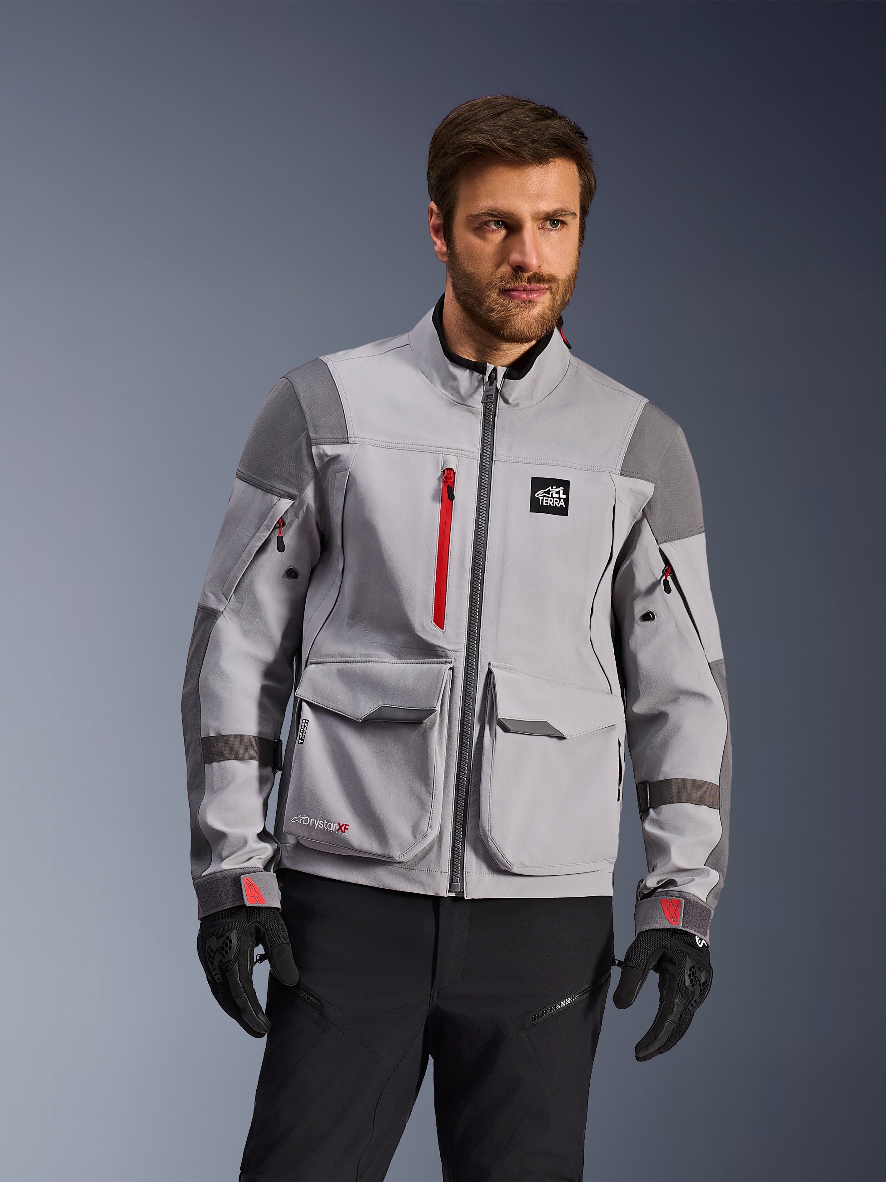 Alpinestars Maxdura Drystar®XF Laminated Armored Shell, chaqueta de motociclismo de aventura, gris medio y gris oscuro con detalles en rojo, vestida por un modelo con guantes y pantalones negros, con grandes bolsillos de carga y cremalleras impermeables, equipamiento profesional para conducción