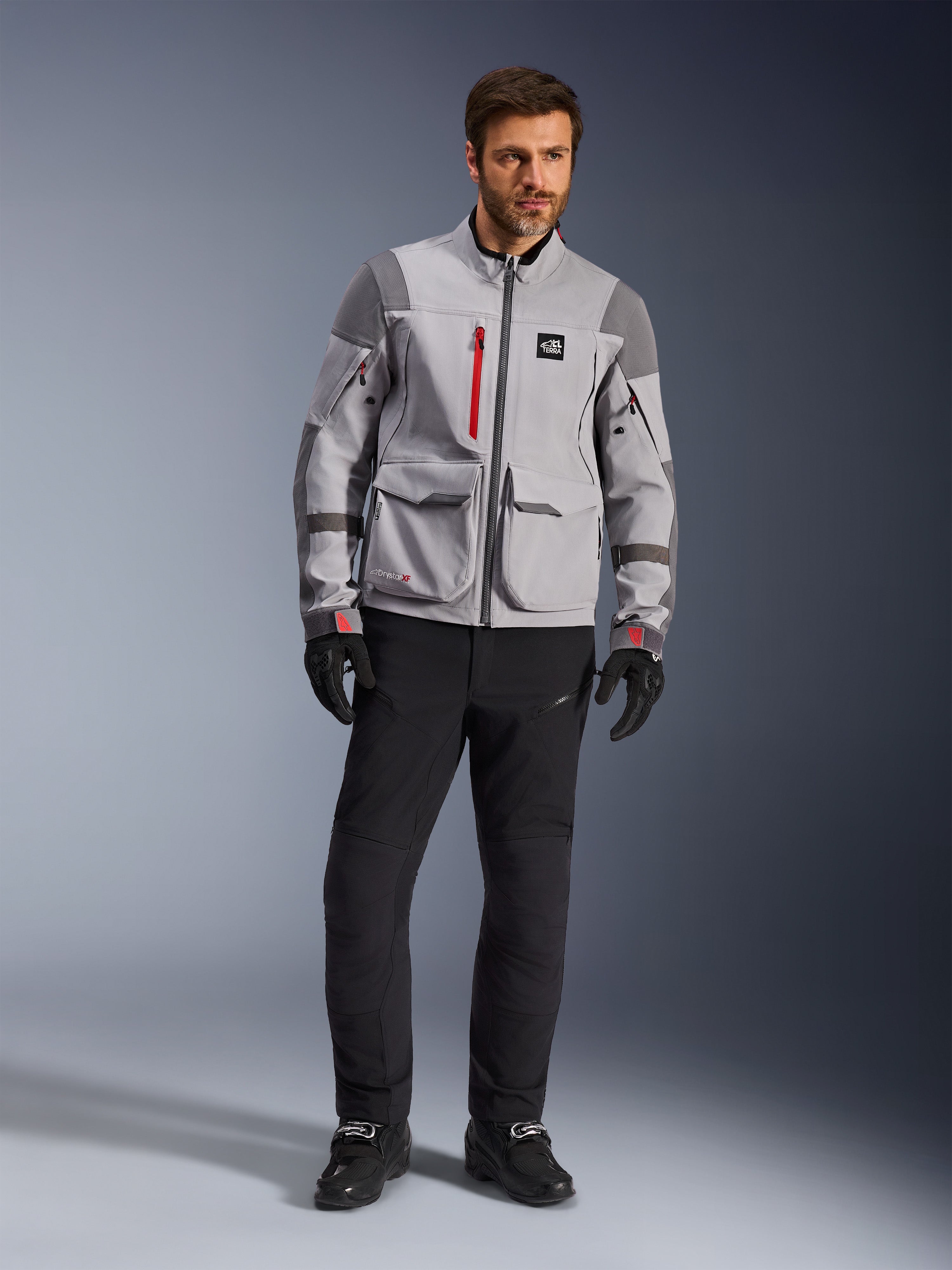 Alpinestars Maxdura Drystar®XF Laminated Armored Shell, chaqueta de aventura para moto, gris medio y gris oscuro con detalles rojos, usada con pantalones y guantes de conducción negros, vista frontal que destaca su construcción técnica y bolsillos de carga.