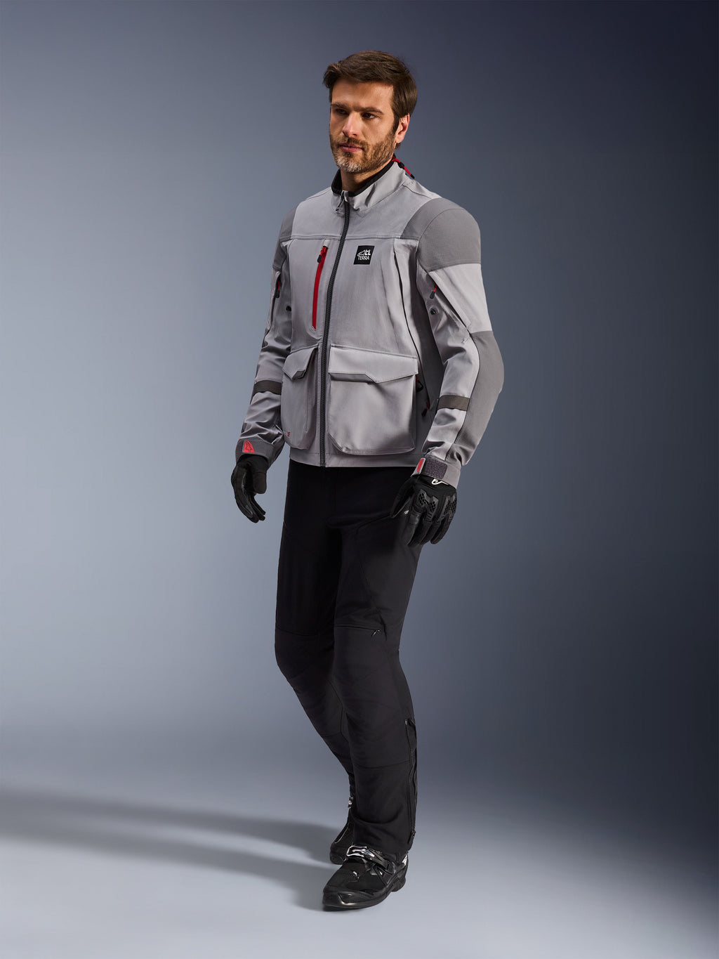 Alpinestars Maxdura Drystar®XF Laminated Armored Shell, chaqueta de motociclismo de aventura, gris medio y gris oscuro, lucida por un modelo con pantalones y guantes de conducción negros, con acentos de cremallera en rojo y bolsillos de carga, equipo técnico profesional para todo clima diseñado para adventure touring.