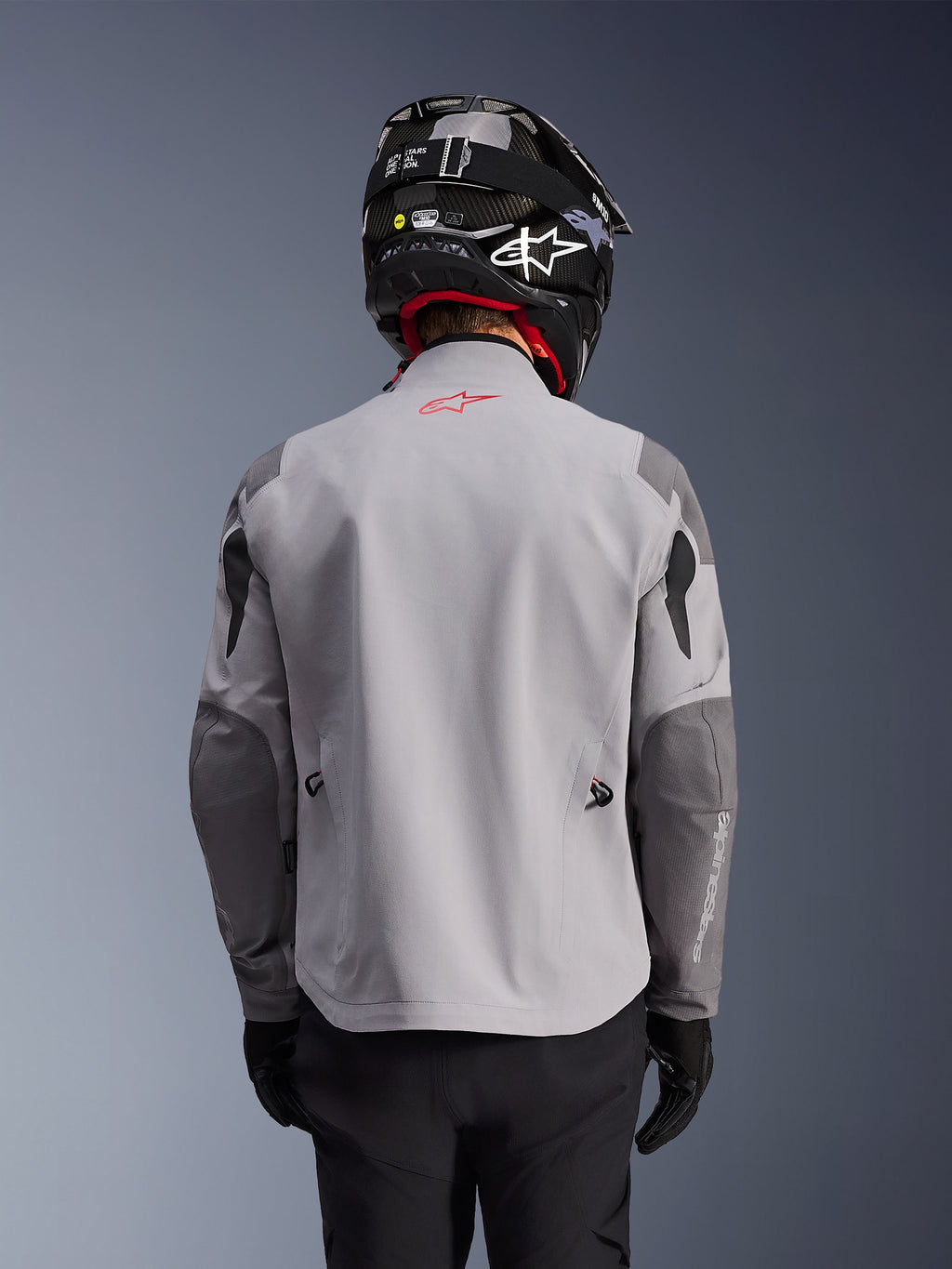 Alpinestars Maxdura Drystar®XF Laminated Armored Shell, chaqueta de moto de aventura, gris medio y gris oscuro, vista trasera que muestra el cuerpo en gris claro con paneles reforzados en gris más oscuro en hombros y codos, logotipo rojo de Alpinestars en la parte superior de la espalda, usada con casco negro y guantes negros.