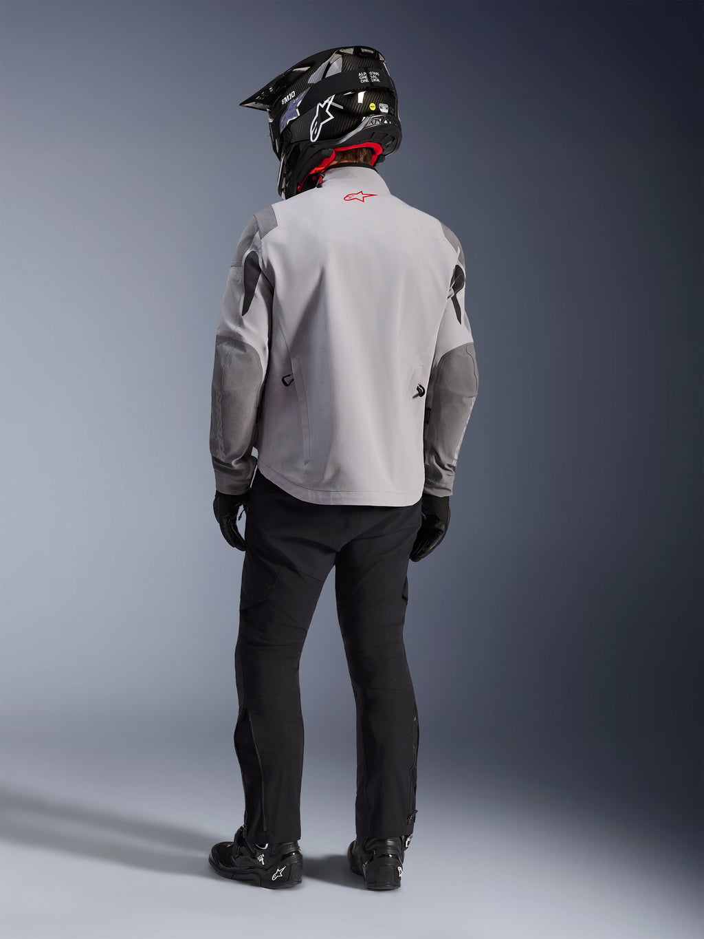 Vista trasera de un motociclista con la chaqueta Alpinestars Maxdura Drystar®XF Laminated Armored Shell en gris medio y gris oscuro, con un logotipo de estrella roja en el cuello, combinada con pantalones de aventura negros, un casco negro con patrón de carbono y botas negras.
