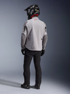 Vista trasera de un motociclista con la chaqueta Alpinestars Maxdura Drystar®XF Laminated Armored Shell en gris medio y gris oscuro, con un logotipo de estrella roja en el cuello, combinada con pantalones de aventura negros, un casco negro con patrón de carbono y botas negras.
