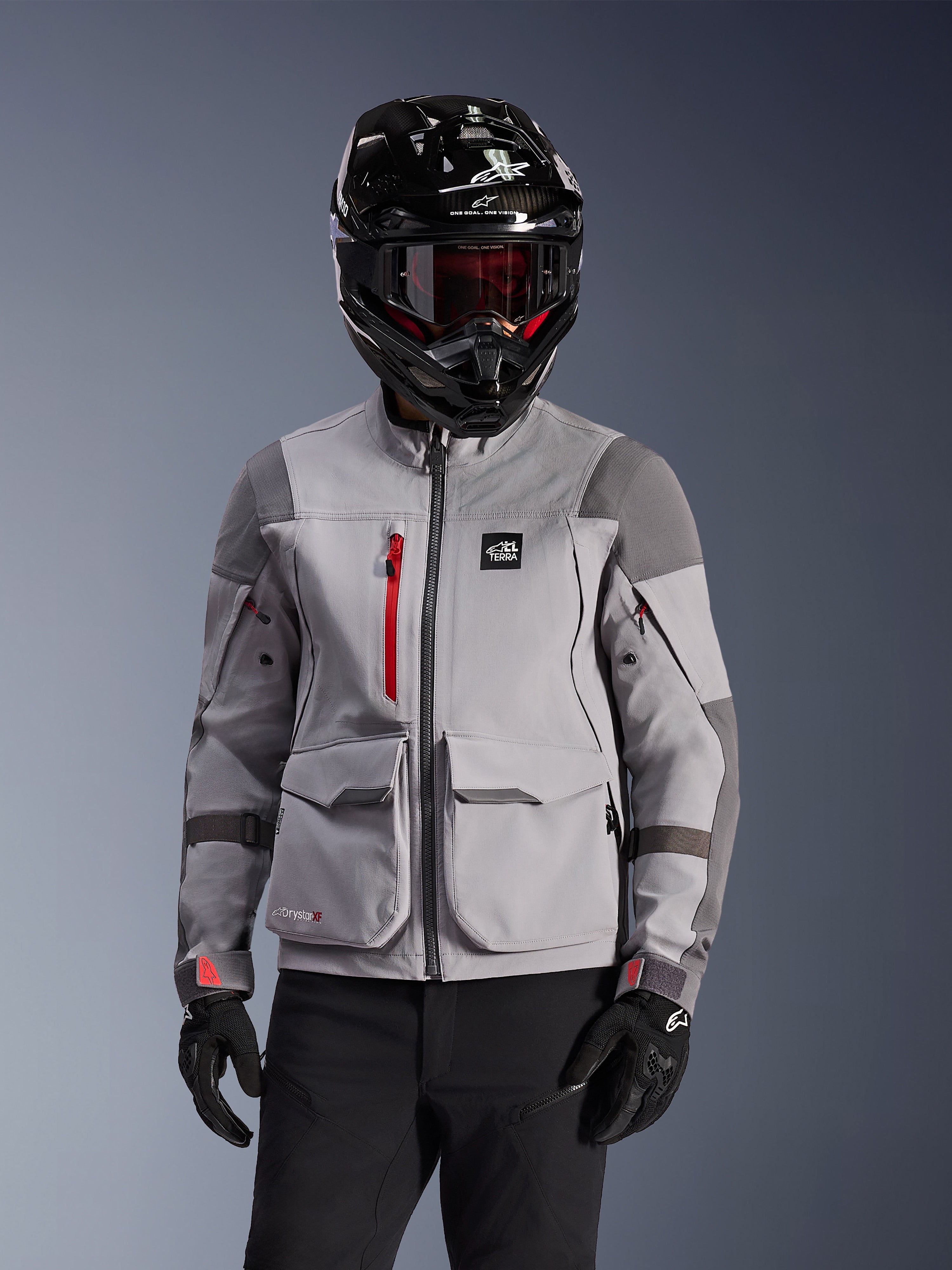 Alpinestars Maxdura Drystar®XF Laminated Armored Shell, chaqueta de motociclismo de aventura en gris medio y gris oscuro, usada con un casco negro de acabado en carbono y gafas de lente roja, con bolsillos de carga frontales y una cremallera roja en el pecho, diseño técnico para adventure touring.