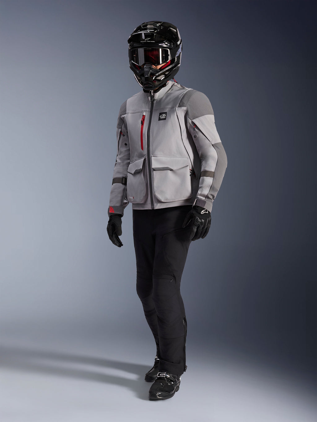 Piloto de aventura con la chaqueta de moto Alpinestars Maxdura Drystar®XF Laminated Armored Shell en gris medio y gris oscuro, casco de aventura negro con gafas, pantalones y guantes negros, con detalles de cremalleras rojas y bolsillos de carga, equipamiento técnico para touring de aventura.