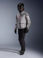 Piloto de aventura con la chaqueta de moto Alpinestars Maxdura Drystar®XF Laminated Armored Shell en gris medio y gris oscuro, casco de aventura negro con gafas, pantalones y guantes negros, con detalles de cremalleras rojas y bolsillos de carga, equipamiento técnico para touring de aventura.