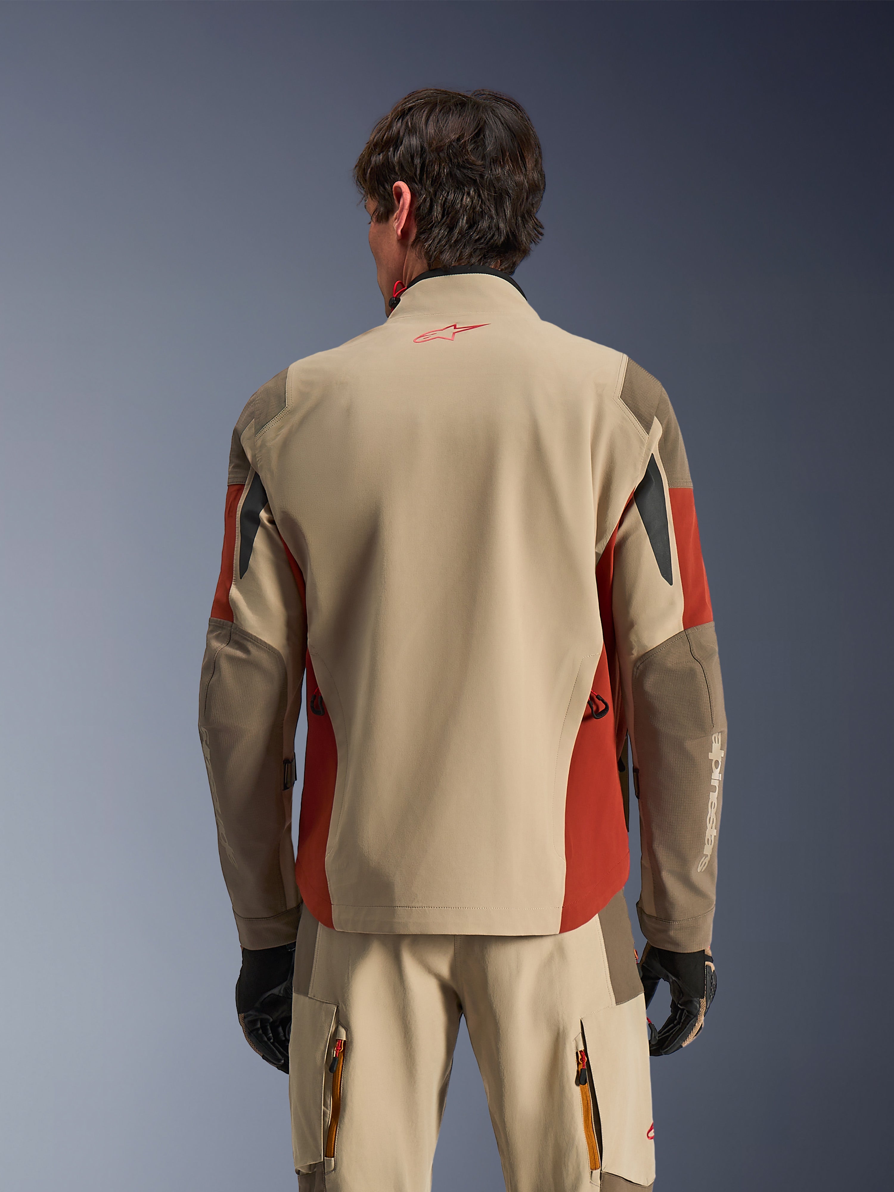 Hombre con la chaqueta Alpinestars Maxdura Drystar®XF Laminated Armored Shell vista desde atrás, chaqueta de aventura en color Aluminum Walnut y Rust Brown, pantalones técnicos a juego y guantes negros, construcción textil laminada de 3 capas con paneles reforzados, conjunto coordinado para motociclismo de aventura