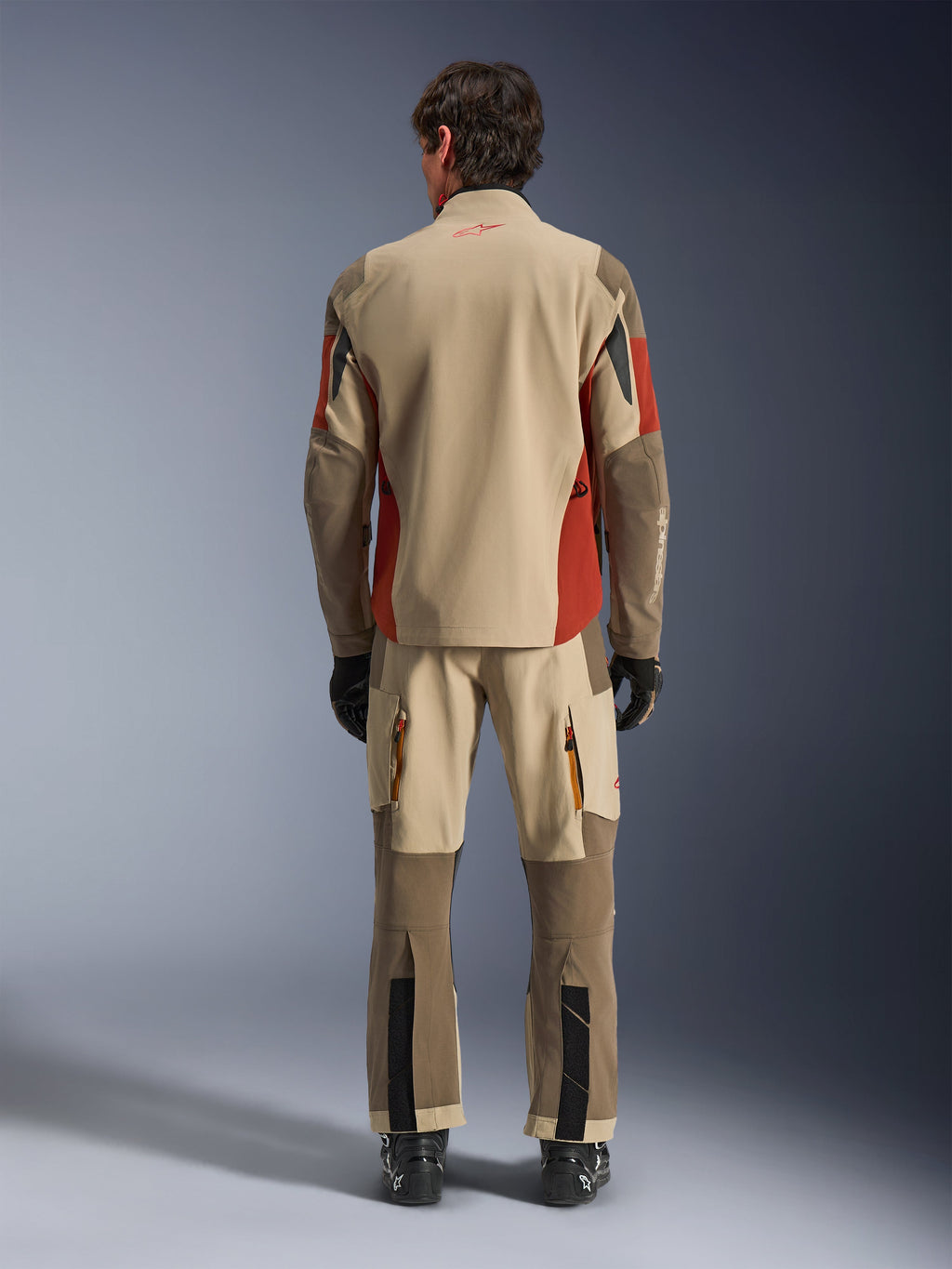 Vista trasera de un piloto con la chaqueta Alpinestars Maxdura Drystar®XF Laminated Armored Shell y pantalones de aventura a juego en Aluminum Walnut Rust Brown, textil canela y marrón con detalles naranjas, combinado con botas y guantes de motociclista negros, equipamiento profesional de adventure touring