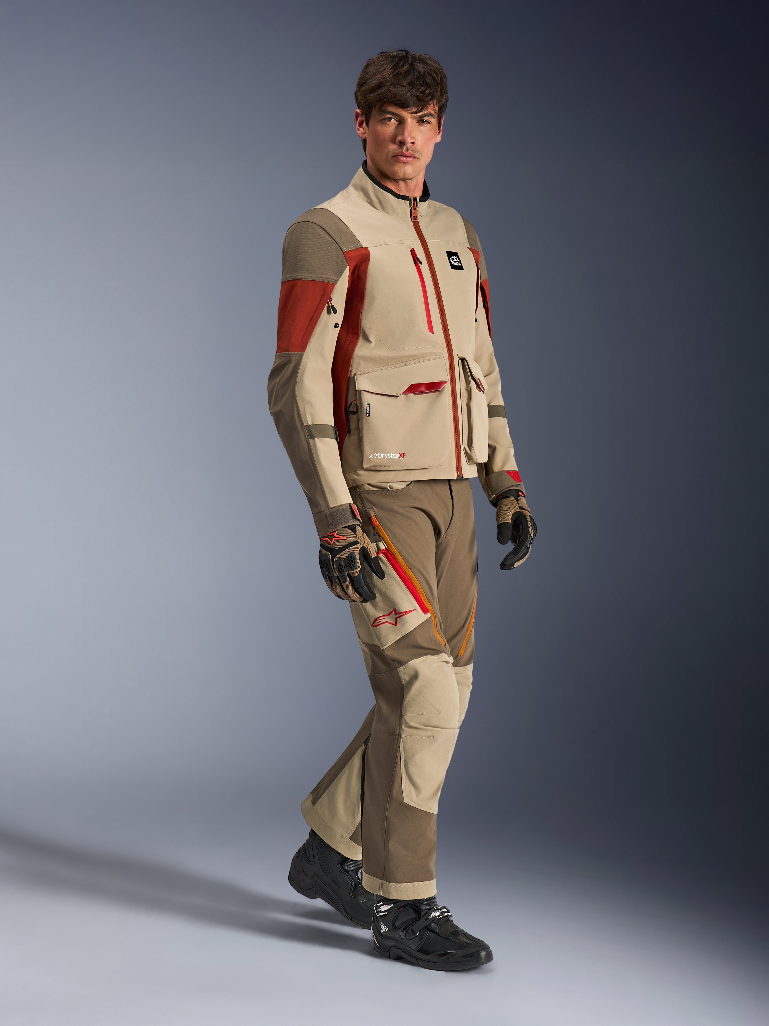 Modelo masculino vistiendo la chaqueta de aventura Alpinestars Maxdura Drystar®XF Laminated Armored Shell en color Aluminum Walnut Rust Brown con pantalones a juego, guantes negros y marrones y botas negras, diseño técnico multicapa con bolsillos de carga y detalles de cremalleras rojas, conjunto coordinado para adventure touring