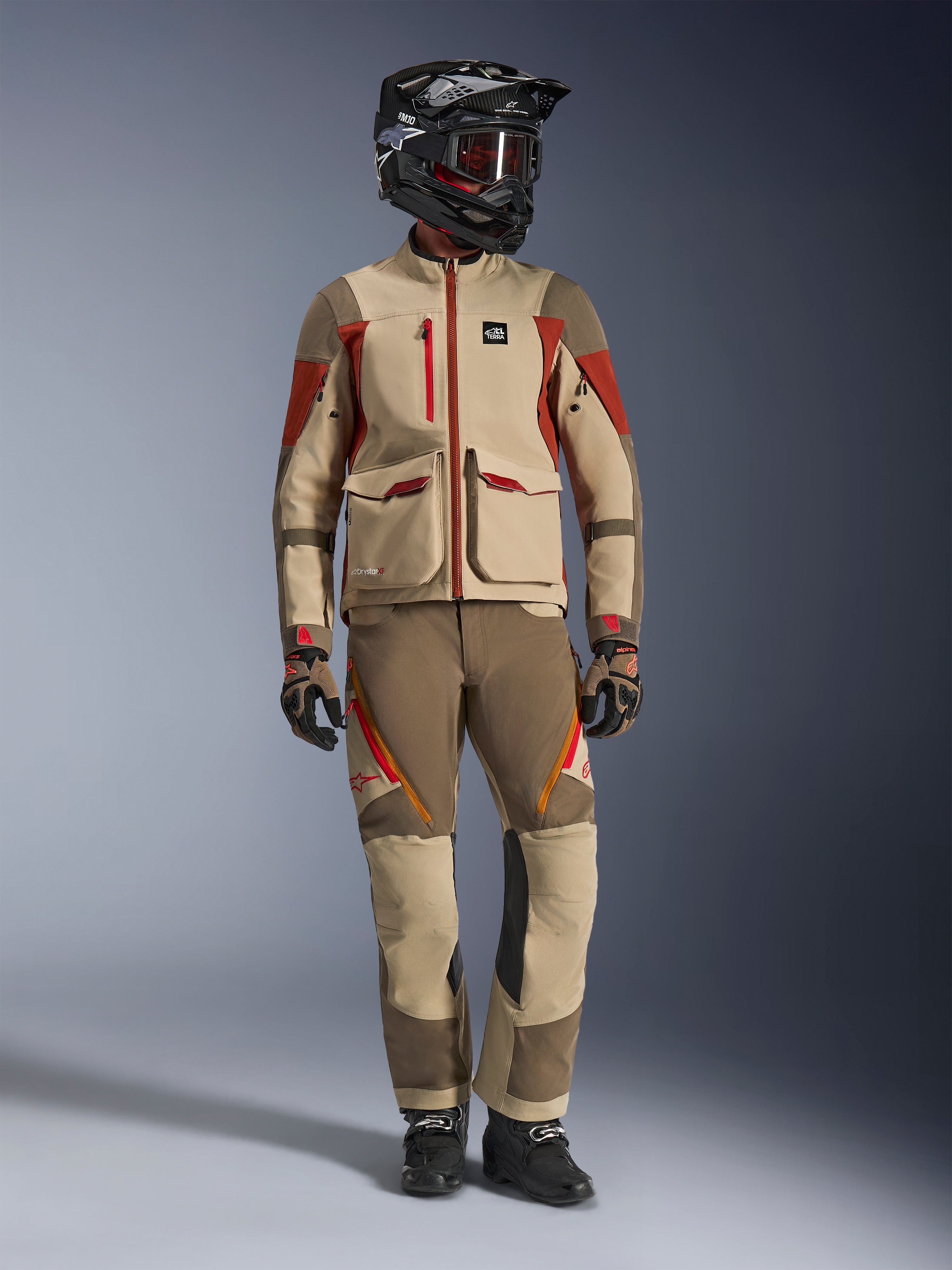 Foto de estudio de cuerpo completo de un piloto con la chaqueta Alpinestars Maxdura Drystar®XF Laminated Armored Shell en Aluminum Walnut Rust Brown, combinada con pantalones de aventura a juego, casco off-road negro con gafas de lentes rojas y guantes técnicos, mostrando un equipamiento completo de adventure touring.