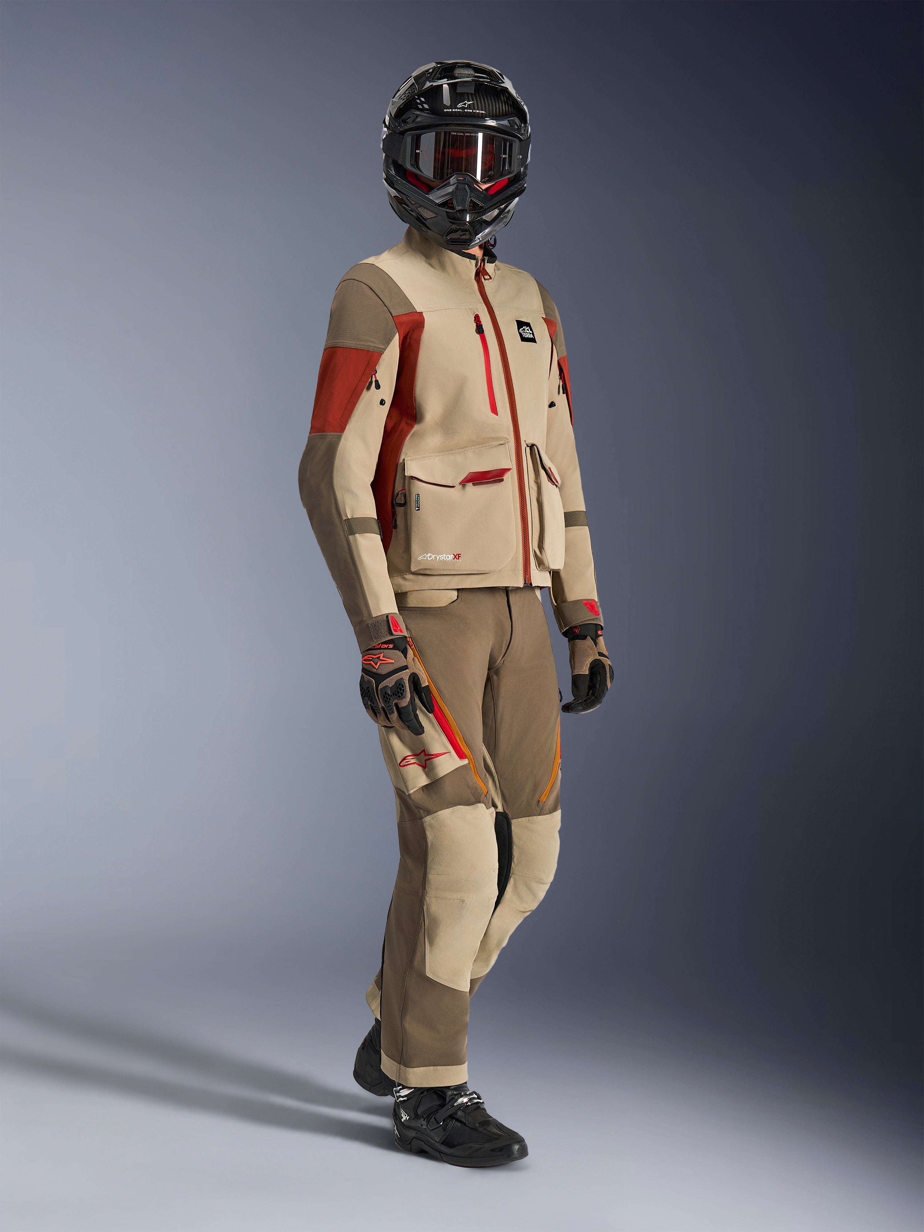 Piloto de aventura vistiendo la chaqueta Maxdura Drystar®XF Laminated Armored Shell en color Aluminum Walnut y Rust Brown con pantalones a juego en nogal y canela, casco negro de fibra de carbono y botas de moto negras, equipamiento técnico de touring para motociclismo de aventura