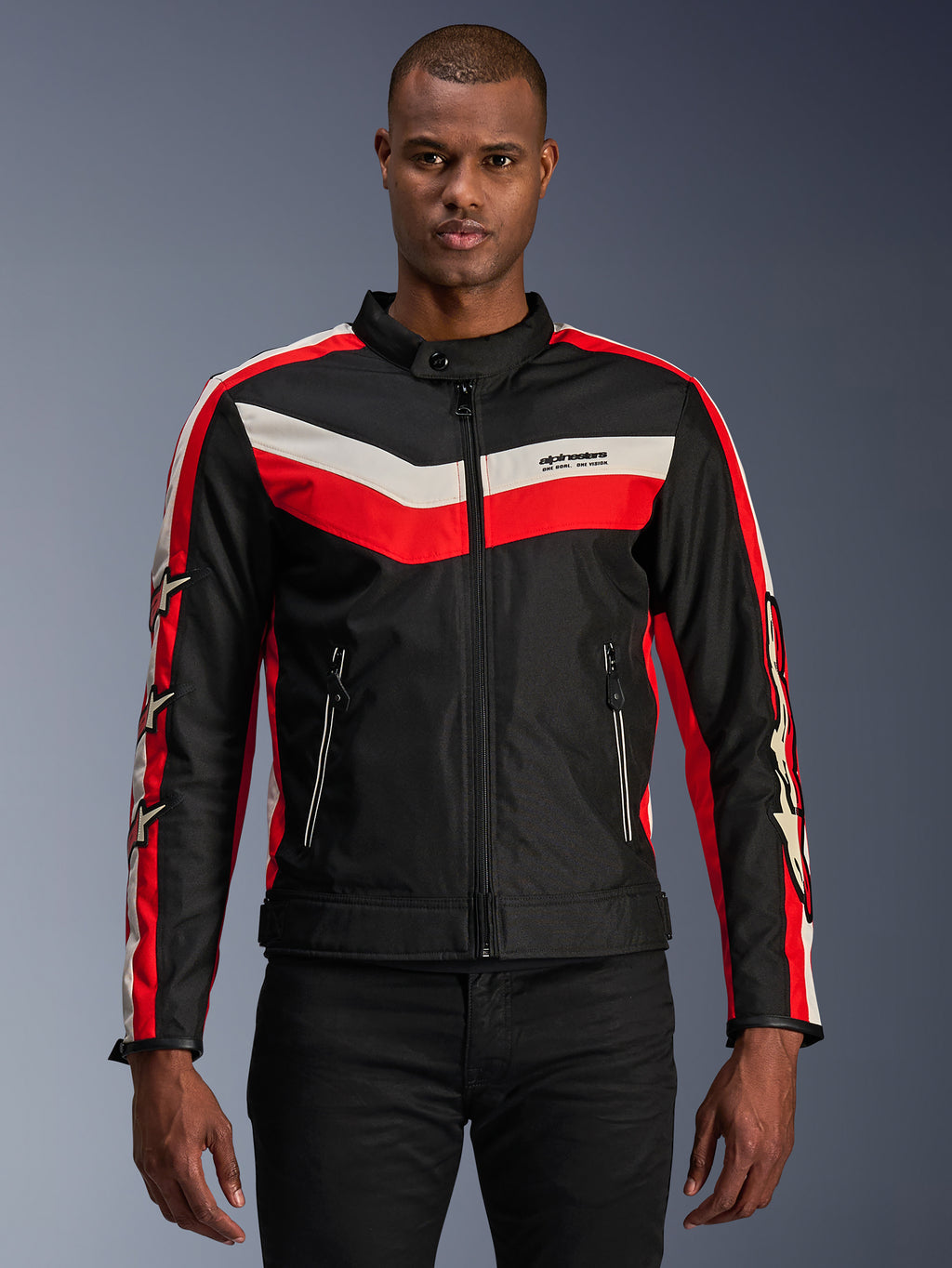 Chaqueta Alpinestars T-Dyno WR, chaqueta textil de moto, negra con franjas tipo chevrón en rojo brillante y crudo, ajuste de motociclista de inspiración vintage, cuello alto con cierre de botón, bolsillos frontales con cremallera, logotipos de estrellas en las mangas, usada por modelo