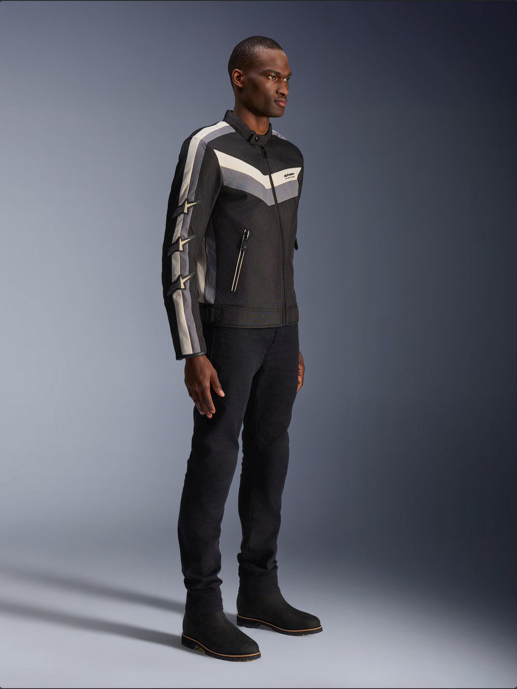 Chaqueta Alpinestars T-Dyno WR, chaqueta textil de moto, negra con acentos de chevrón en gris oscuro y crudo, ajuste de motociclista de inspiración vintage, combinada con pantalones y botas negras para un look completo de carretera