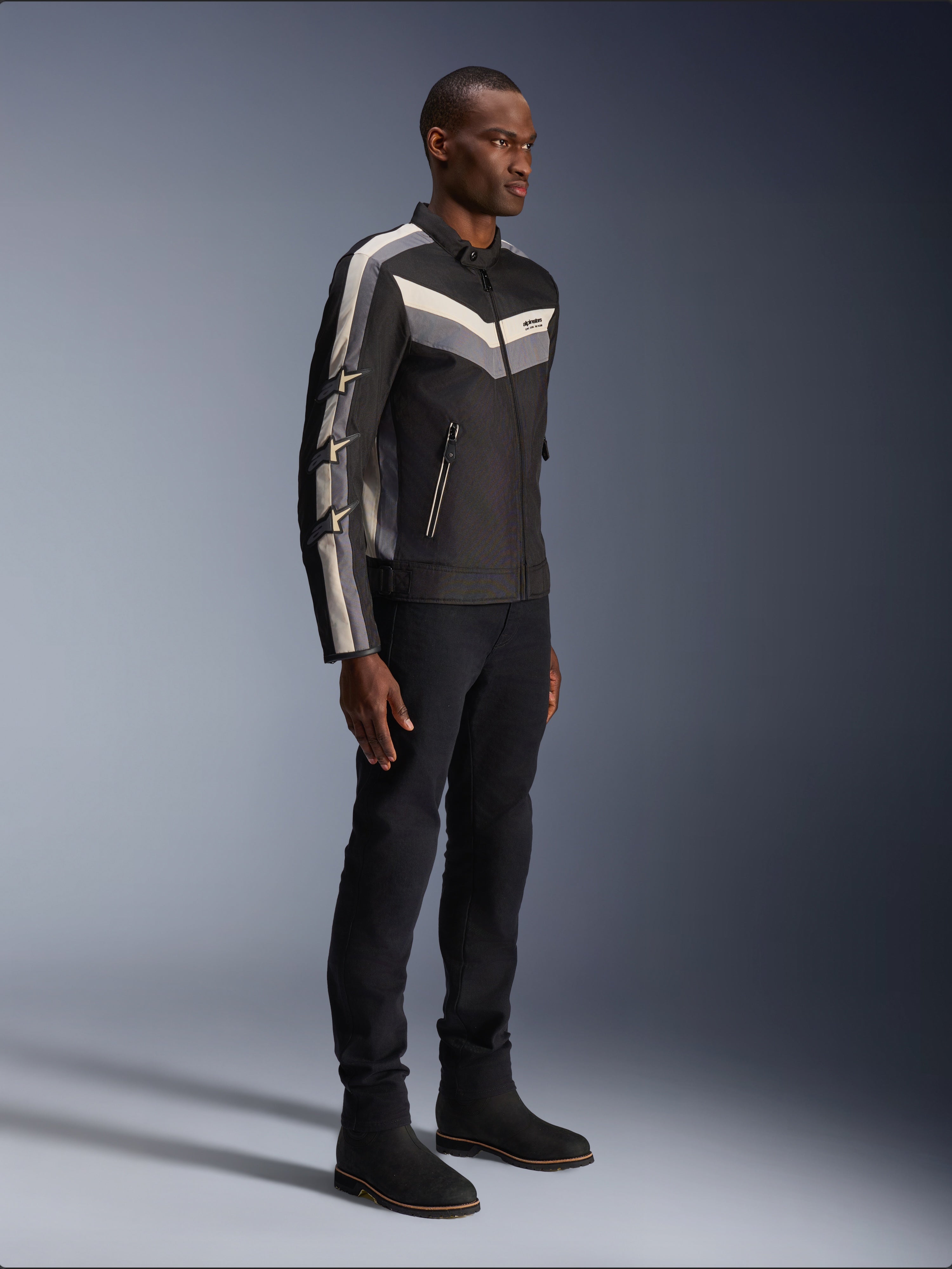Chaqueta Alpinestars T-Dyno WR, chaqueta textil de moto, negra con acentos de chevrón en gris oscuro y crudo, ajuste de motociclista de inspiración vintage, combinada con pantalones y botas negras para un look completo de carretera