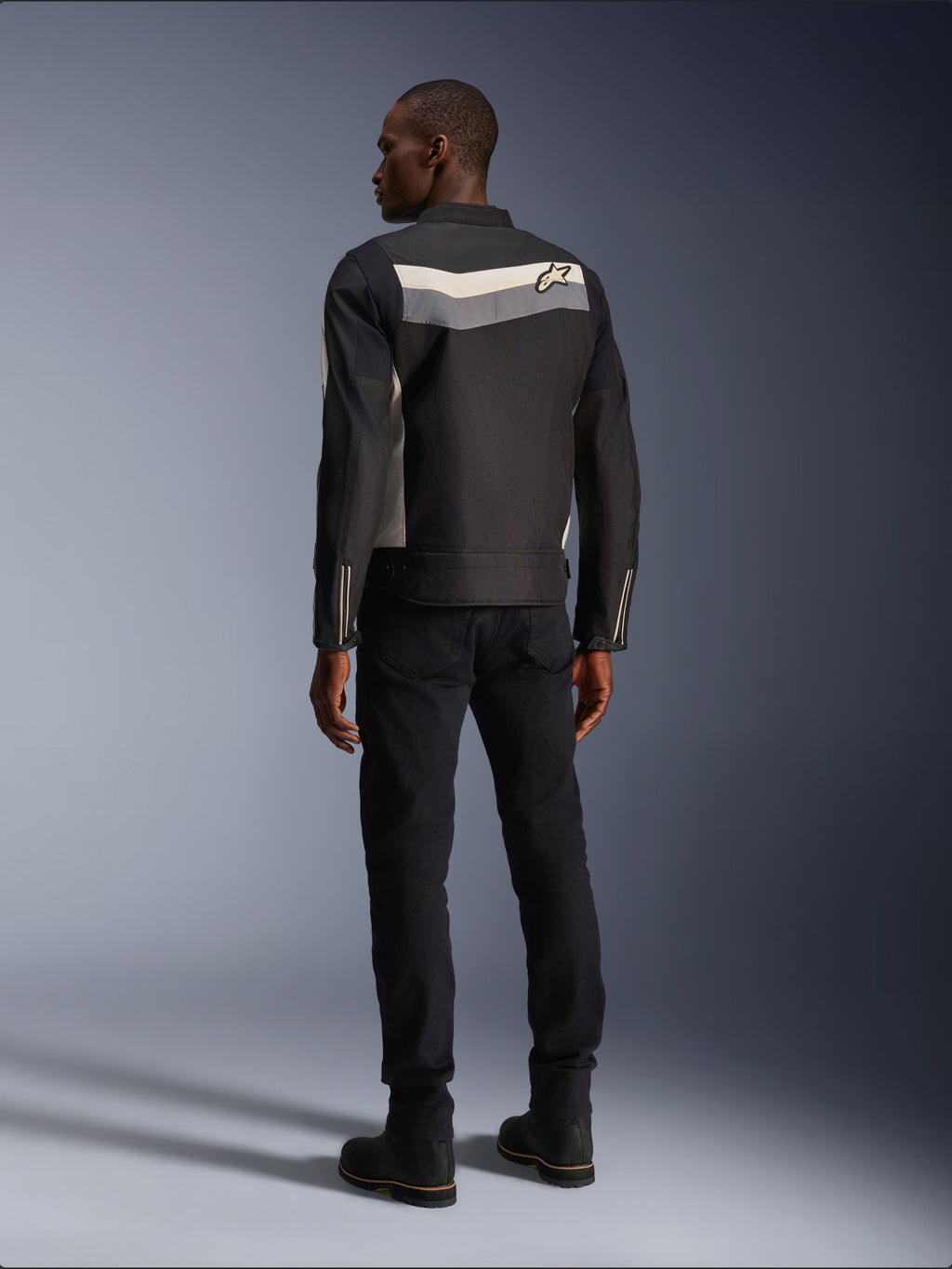 Modelo masculino de espaldas vistiendo la chaqueta Alpinestars T-Dyno WR en color negro, gris oscuro y crudo, chaqueta textil de moto con una franja horizontal y el logo de la estrella en la espalda, combinada con pantalones y botas de moto negros, estética de equipamiento urbano para motociclista.