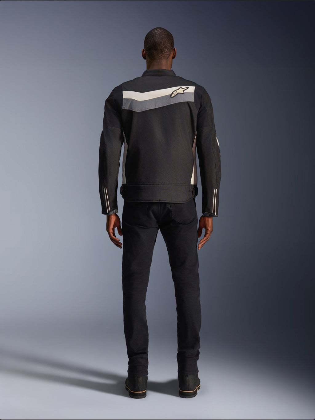 Chaqueta Alpinestars T-Dyno WR, chaqueta de moto textil, Negro Gris Oscuro y Crudo, vista posterior que muestra una franja horizontal en los hombros con un pequeño logotipo de estrella, usada por un modelo con pantalones y botas negros, equipamiento de carretera de inspiración vintage