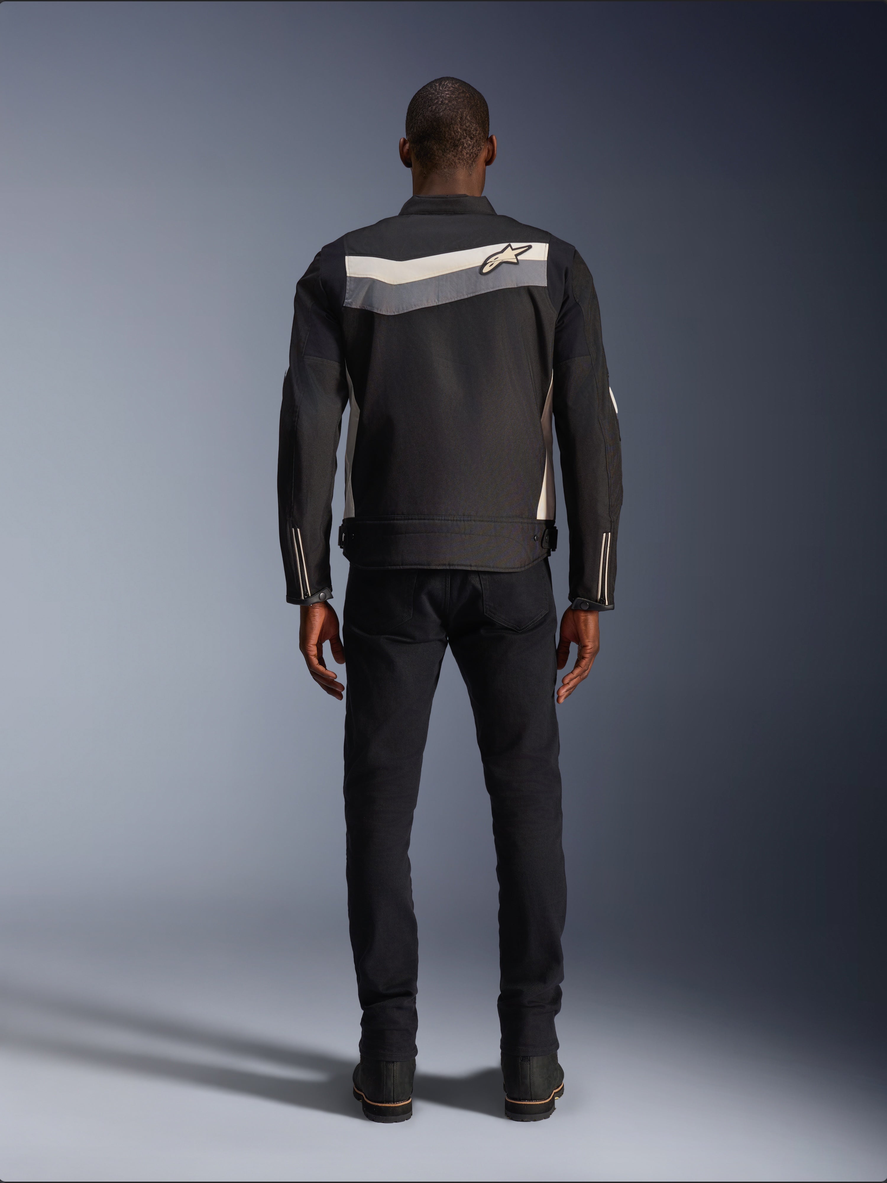 Chaqueta Alpinestars T-Dyno WR, chaqueta de moto textil, Negro Gris Oscuro y Crudo, vista posterior que muestra una franja horizontal en los hombros con un pequeño logotipo de estrella, usada por un modelo con pantalones y botas negros, equipamiento de carretera de inspiración vintage
