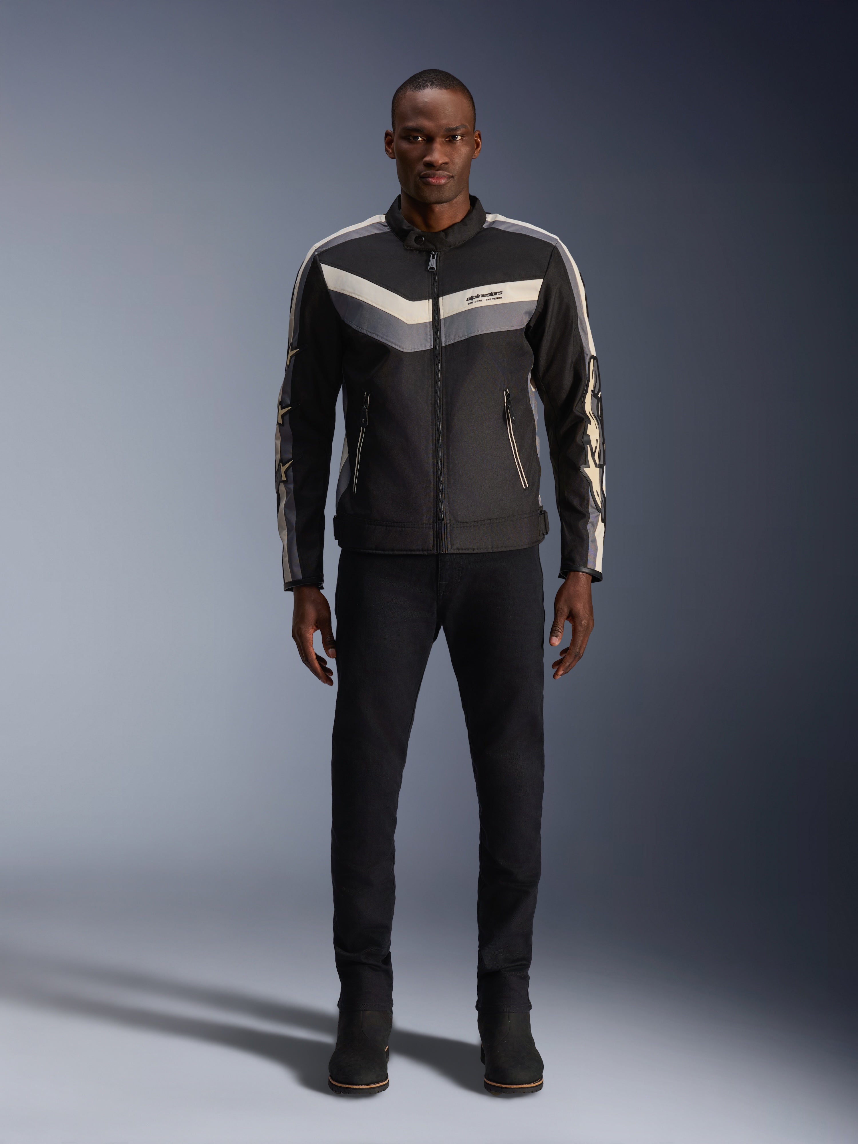 Chaqueta Alpinestars T-Dyno WR, negro, gris oscuro y crudo, chaqueta de moto textil con patrón de chevrón en el pecho de inspiración vintage, usada por modelo masculino con pantalones y botas de montar negros, equipamiento de carretera urbano contemporáneo