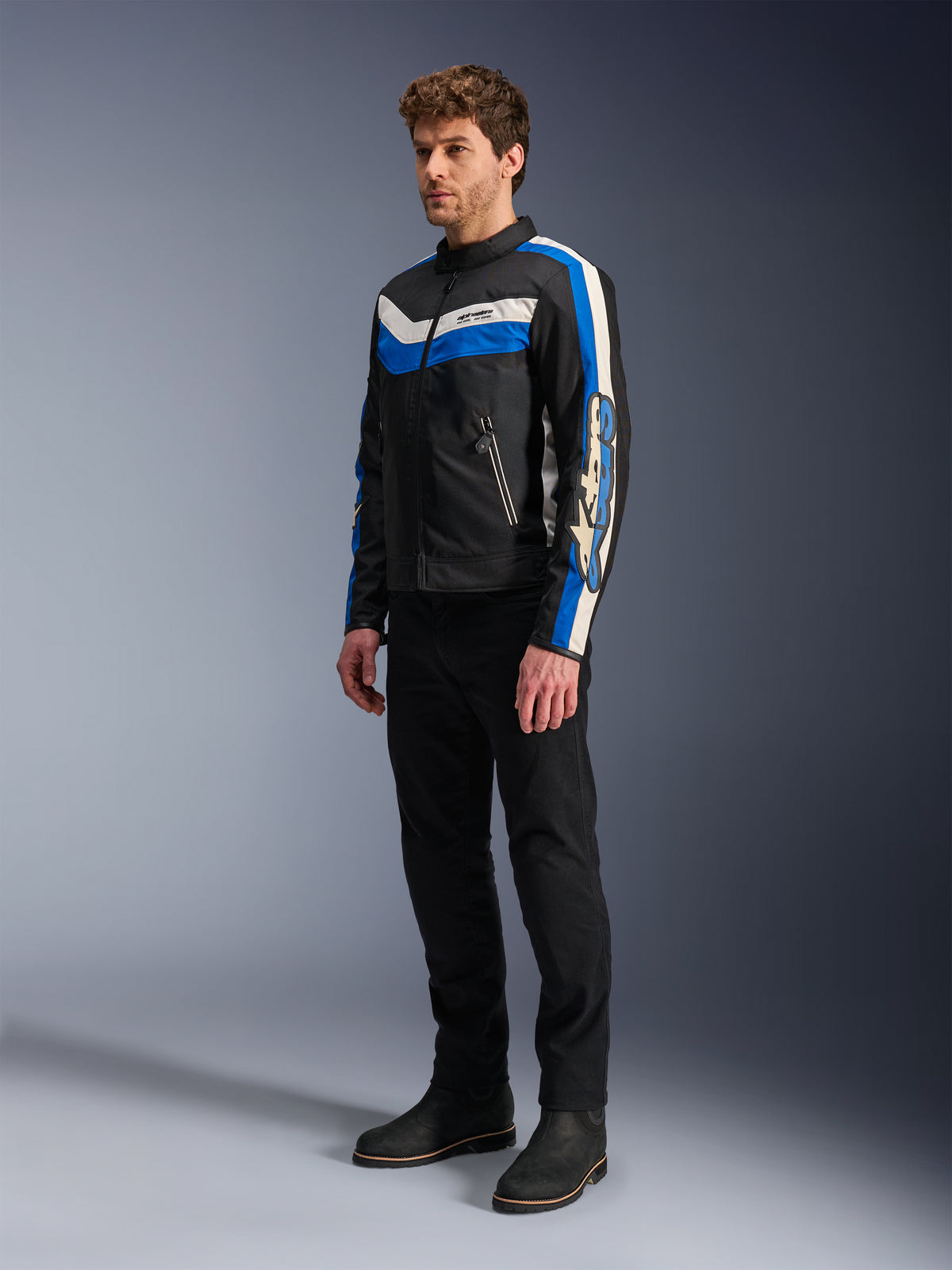 Chaqueta Alpinestars T-Dyno WR, negro, azul y crudo, chaqueta textil de moto usada por un modelo con pantalones y botas negros, diseño de inspiración vintage con llamativas rayas azules y crudo y logotipos en las mangas, fotografía de estudio