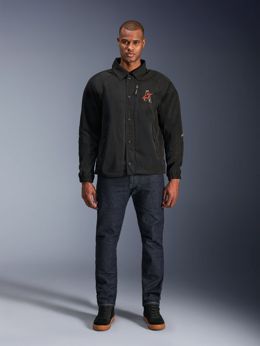 Chaqueta Alpinestars Revved WR en color negro, vestida por un modelo con vaqueros azul oscuro y zapatillas negras, vista frontal, con cierre de botones a presión y logotipo bordado en 3D en el pecho, equipamiento de motociclismo urbano contemporáneo.